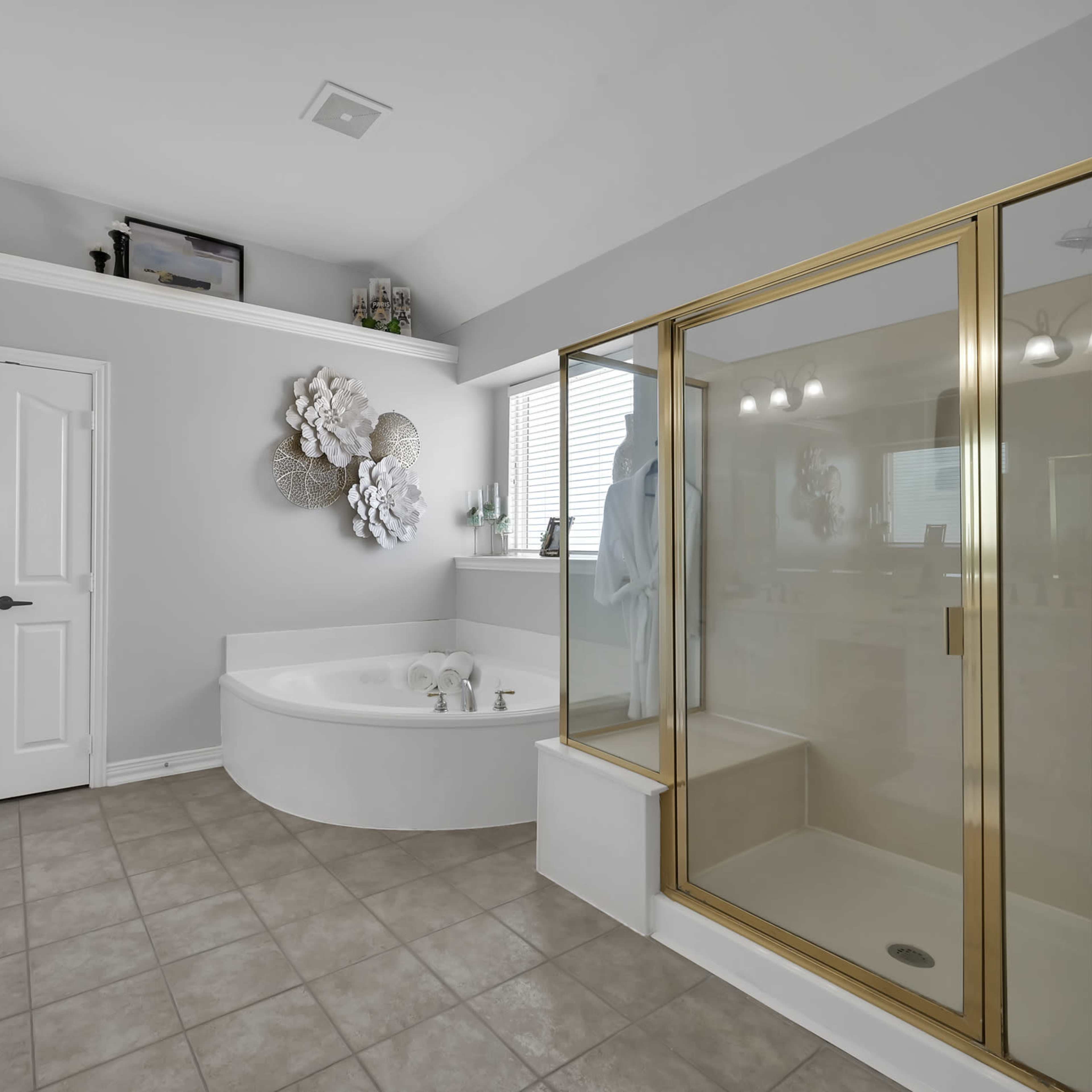 The bathroom features a curved soaking tub beside a glass-enclosed shower with gold trim, against light gray walls and tiled flooring.