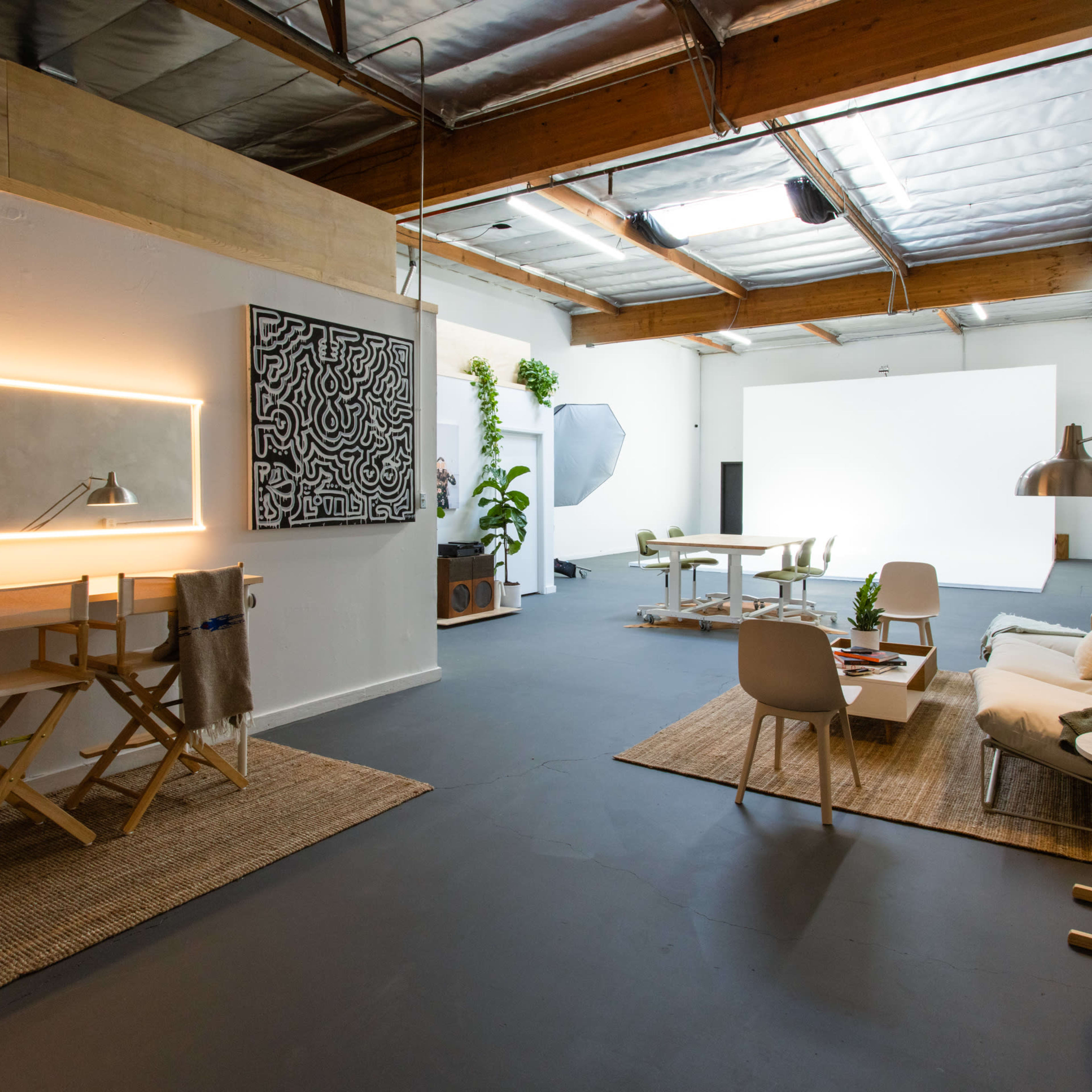 A spacious, well-lit studio featuring a seating area, a workspace with a desk and lamp, and a large blank wall suitable for presentations or photography.