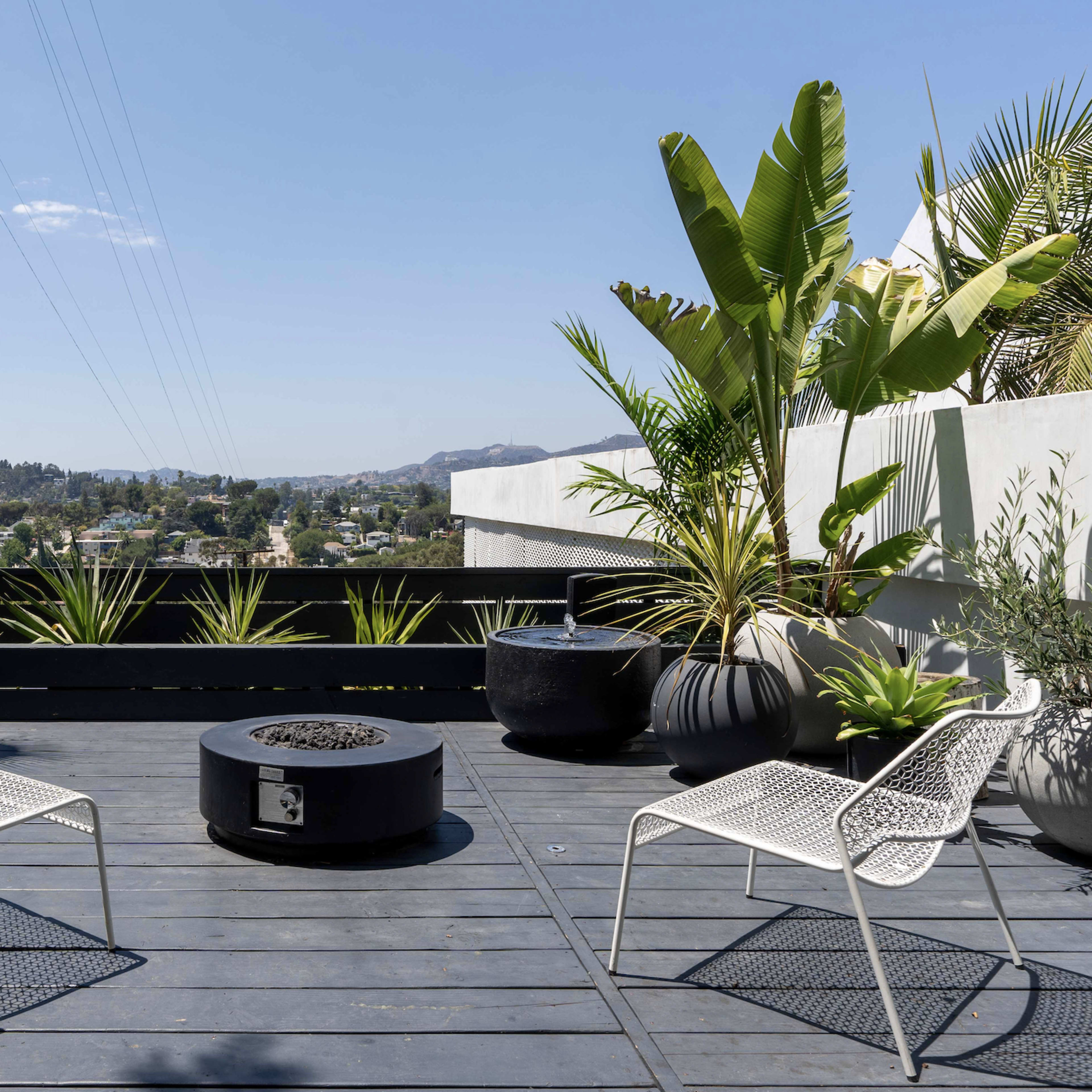 A modern rooftop terrace features two white metal chairs arranged around a circular fire pit, surrounded by potted plants and a scenic view of the town below.