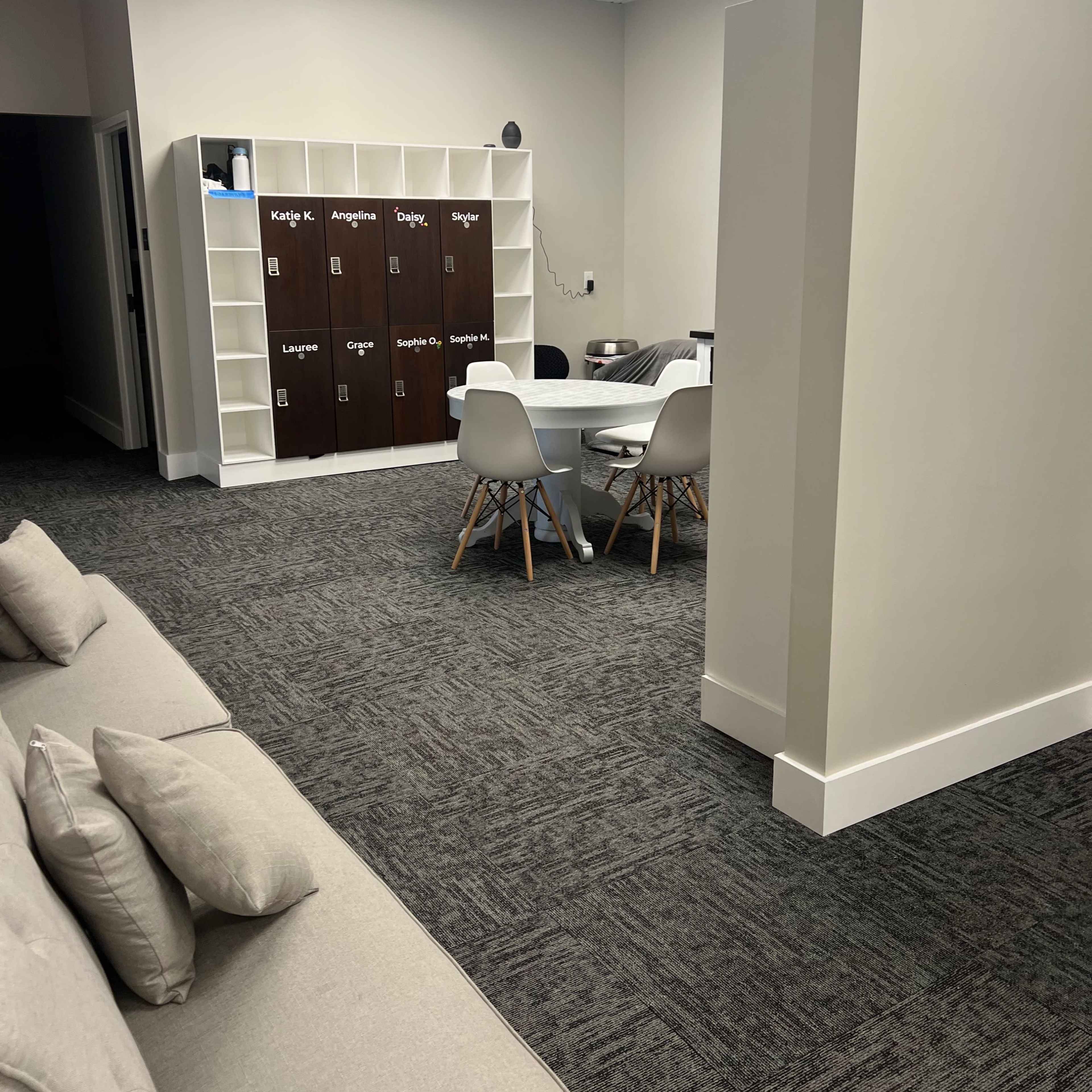 A modern office space with a seating area featuring a light-colored couch and a workspace that includes a table surrounded by four chairs and a row of dark brown lockers against the wall.