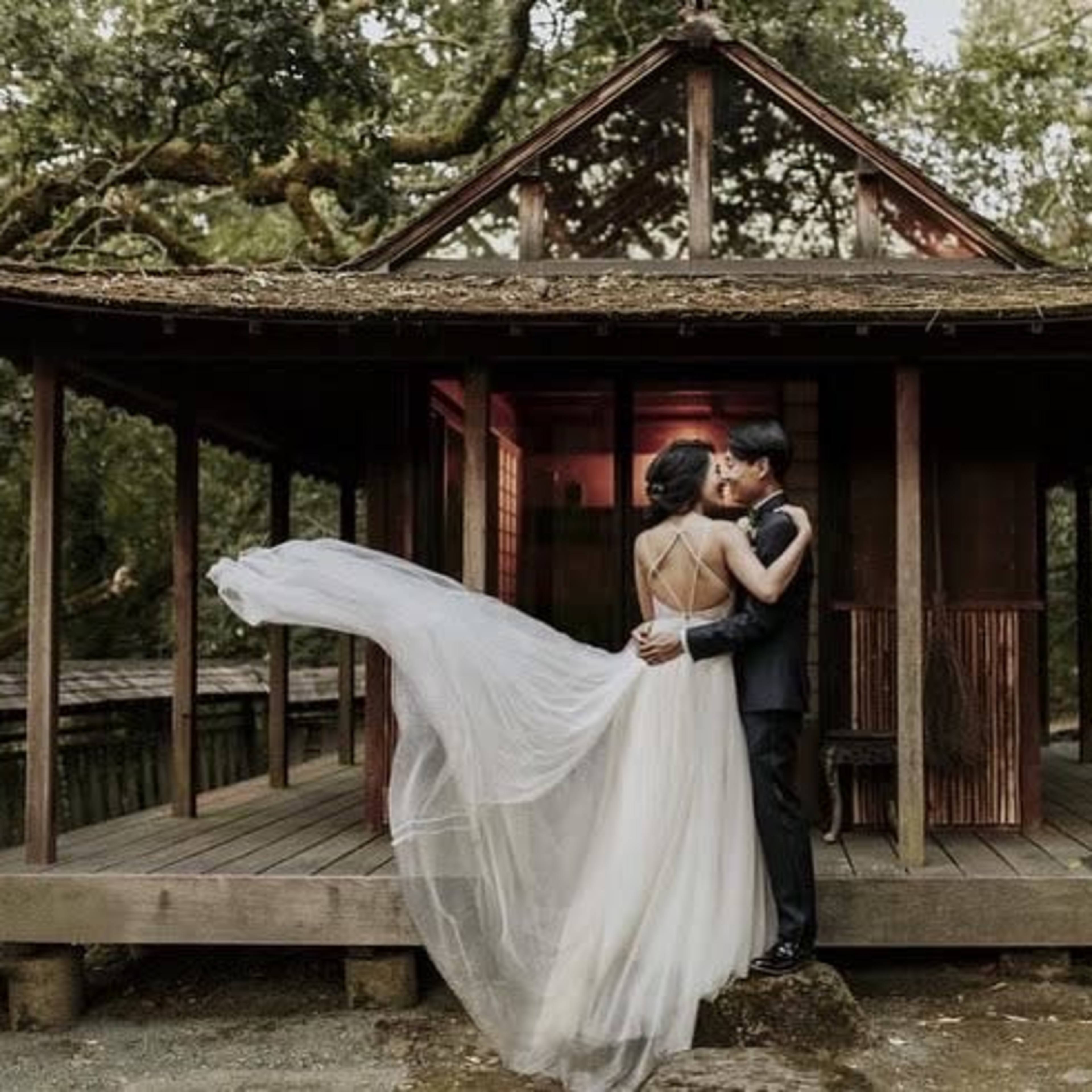 A couple embraces in front of a traditional wooden structure surrounded by trees and a pond.