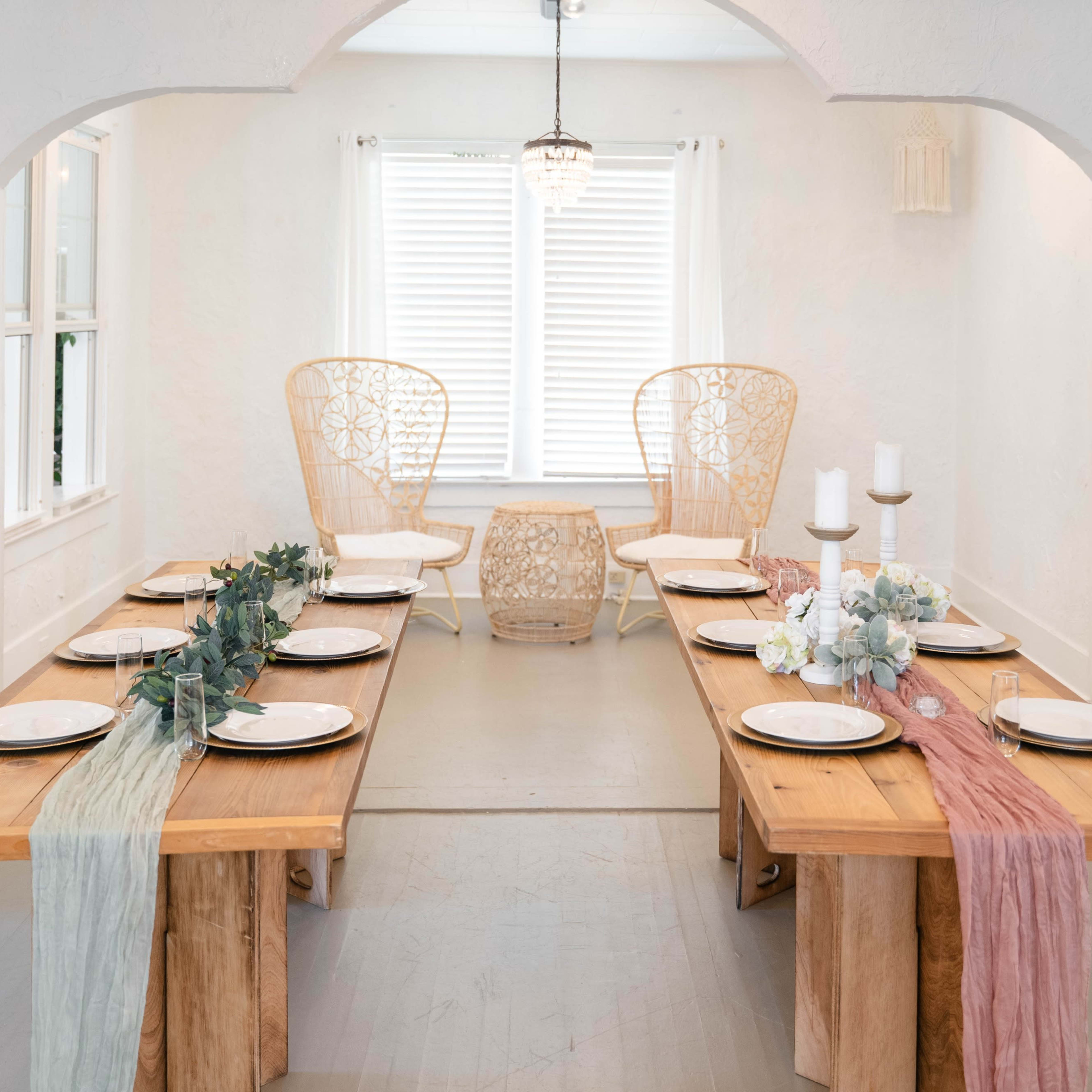 A minimalist dining room features two long wooden tables set with plates, surrounded by light-colored walls and framed by decorative chairs and a small table near a window.