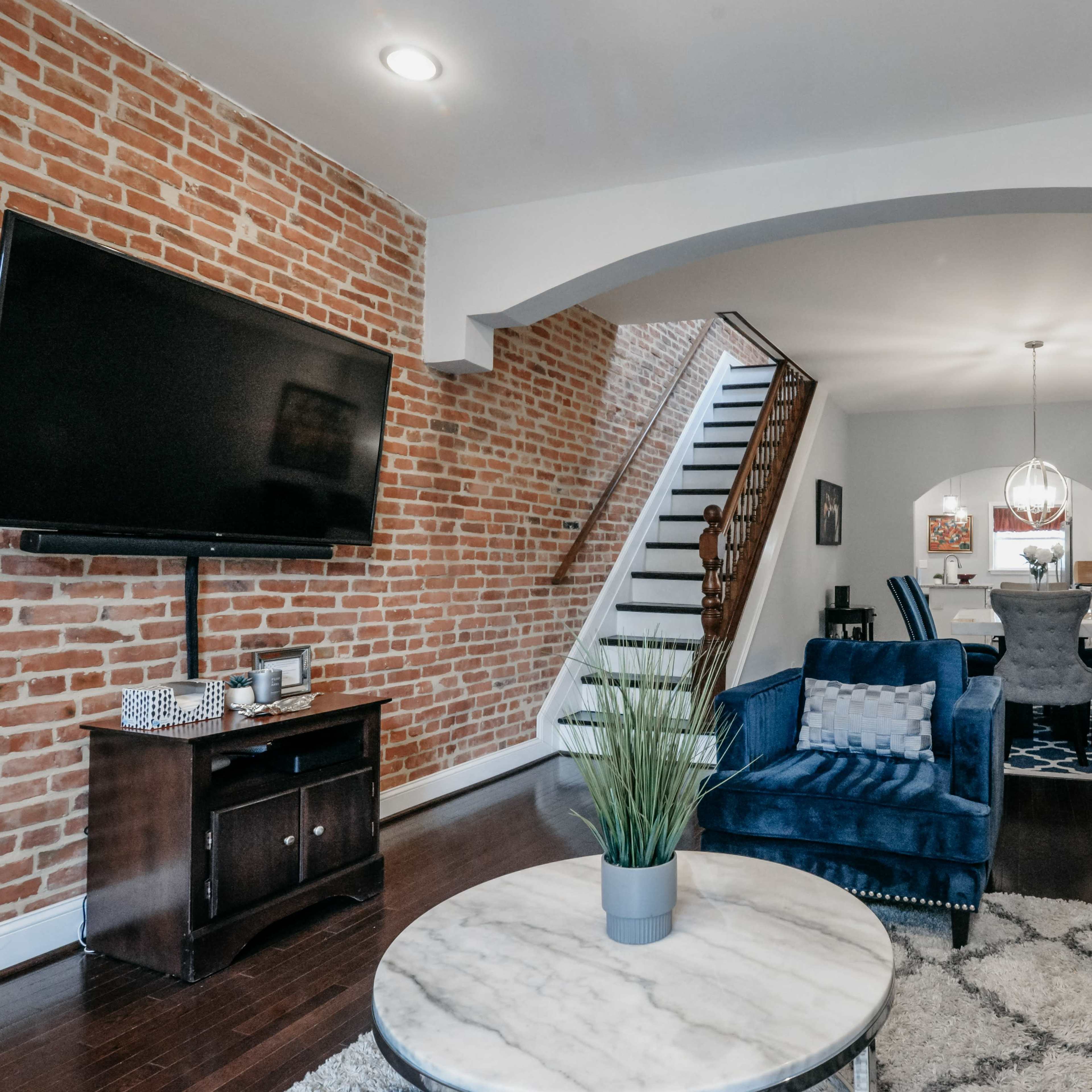 A stylish living room with exposed brick walls, a large TV mounted on the wall, and a seating area featuring a blue armchair and a gray sofa, leading to a dining area with a table and chairs.