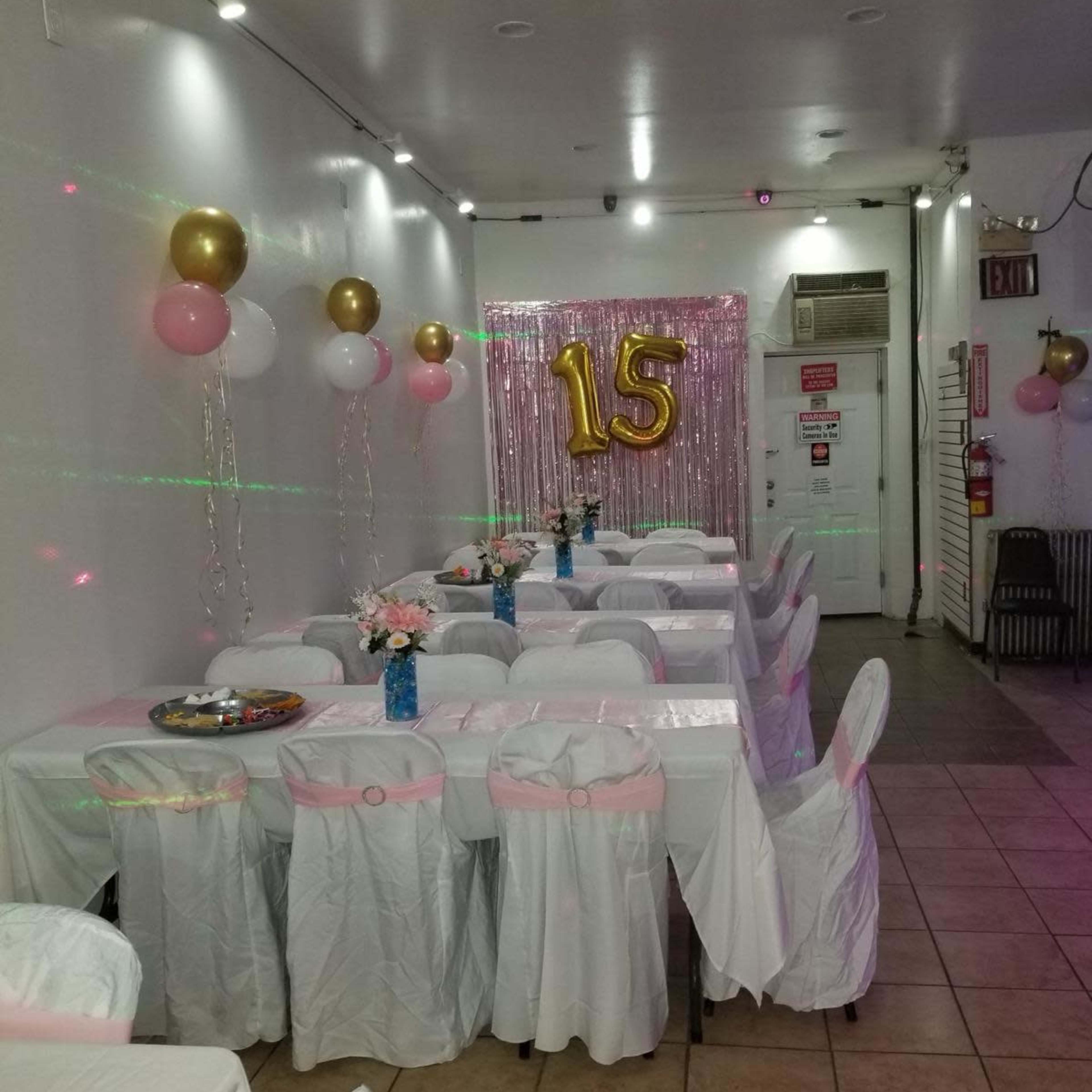 A decorated event space with tables covered in white tablecloths, pink and gold balloons, and a large "15" displayed against a shimmering backdrop.