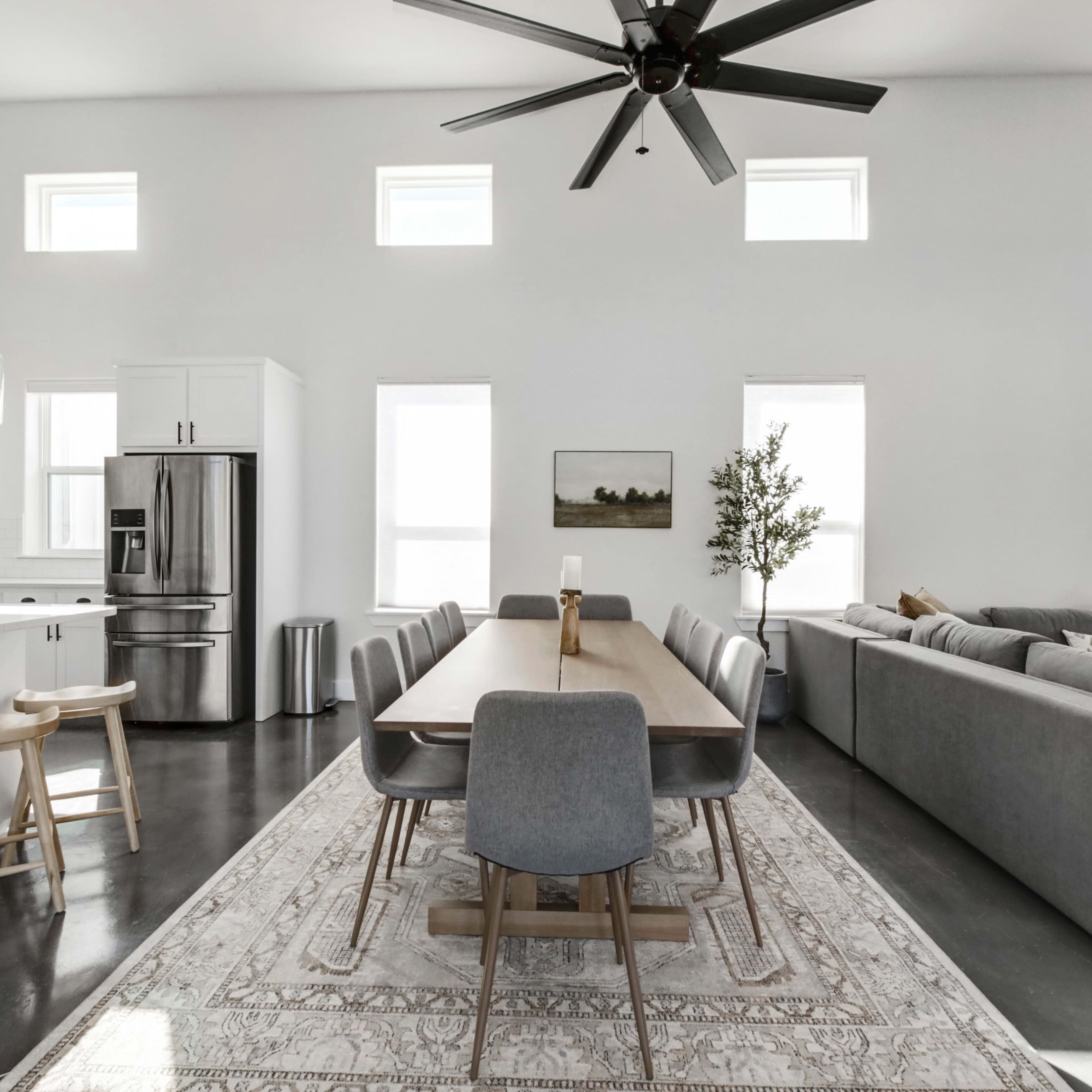 A spacious, modern living area with a dining table, chairs, a kitchen area, and a large sectional sofa, all arranged under a high ceiling with large windows.