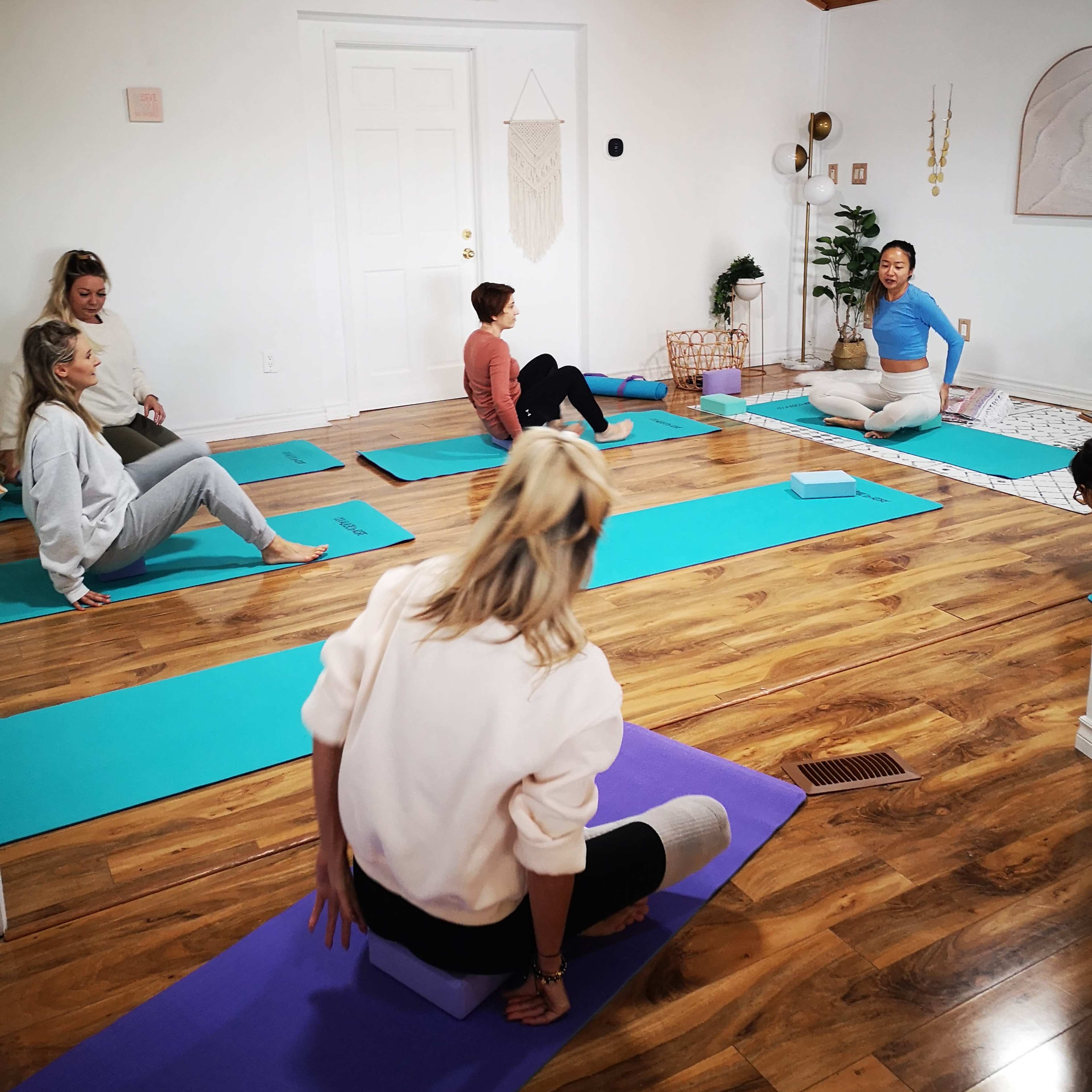 A group of people is seated on yoga mats in a well-lit room, listening to an instructor who is sitting on the floor in front of them.