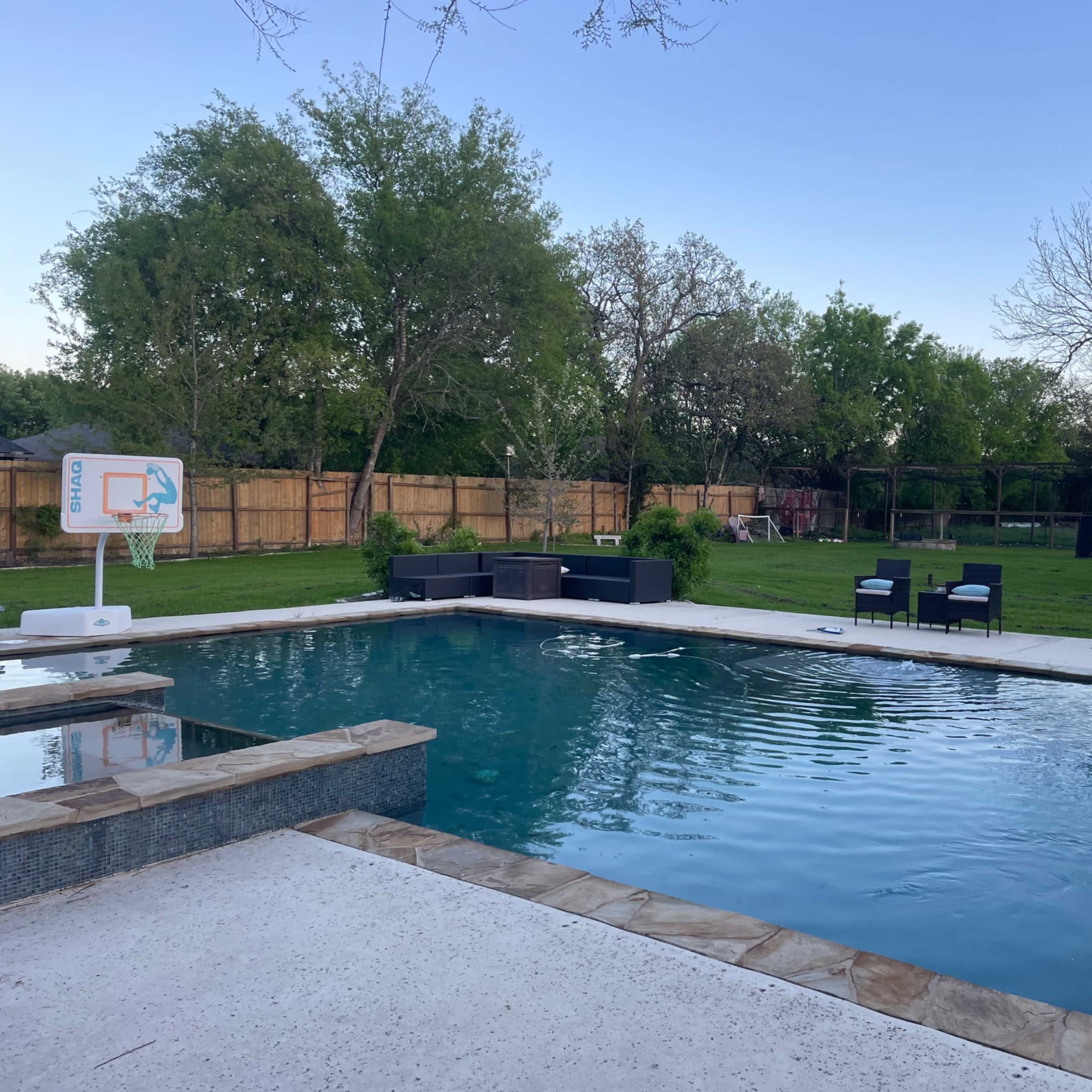A large rectangular swimming pool is surrounded by a stone patio and green grass, with black lounge chairs positioned nearby.