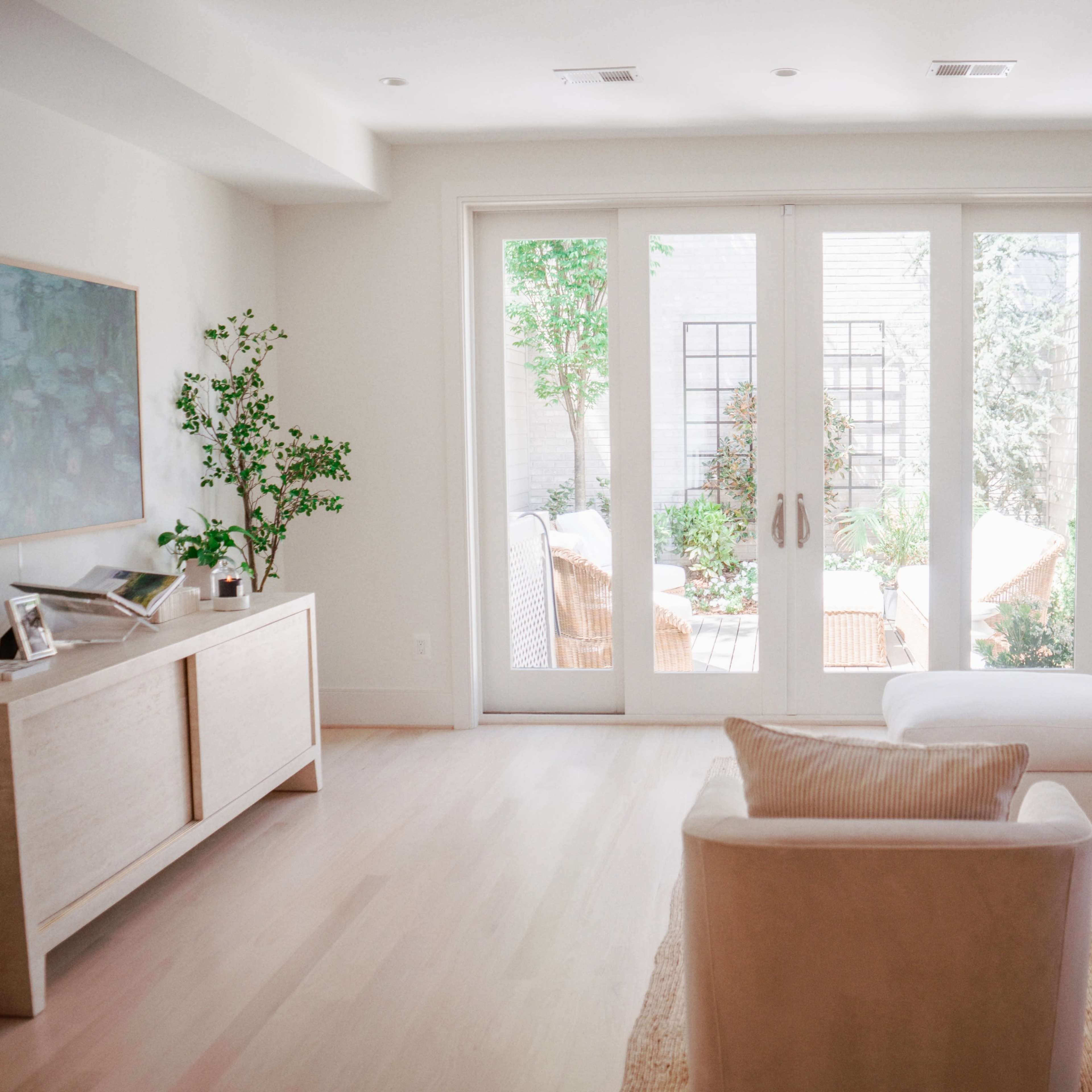 A bright living room features a white sectional sofa, a light wooden media console, and large glass doors that open to a landscaped outdoor area.