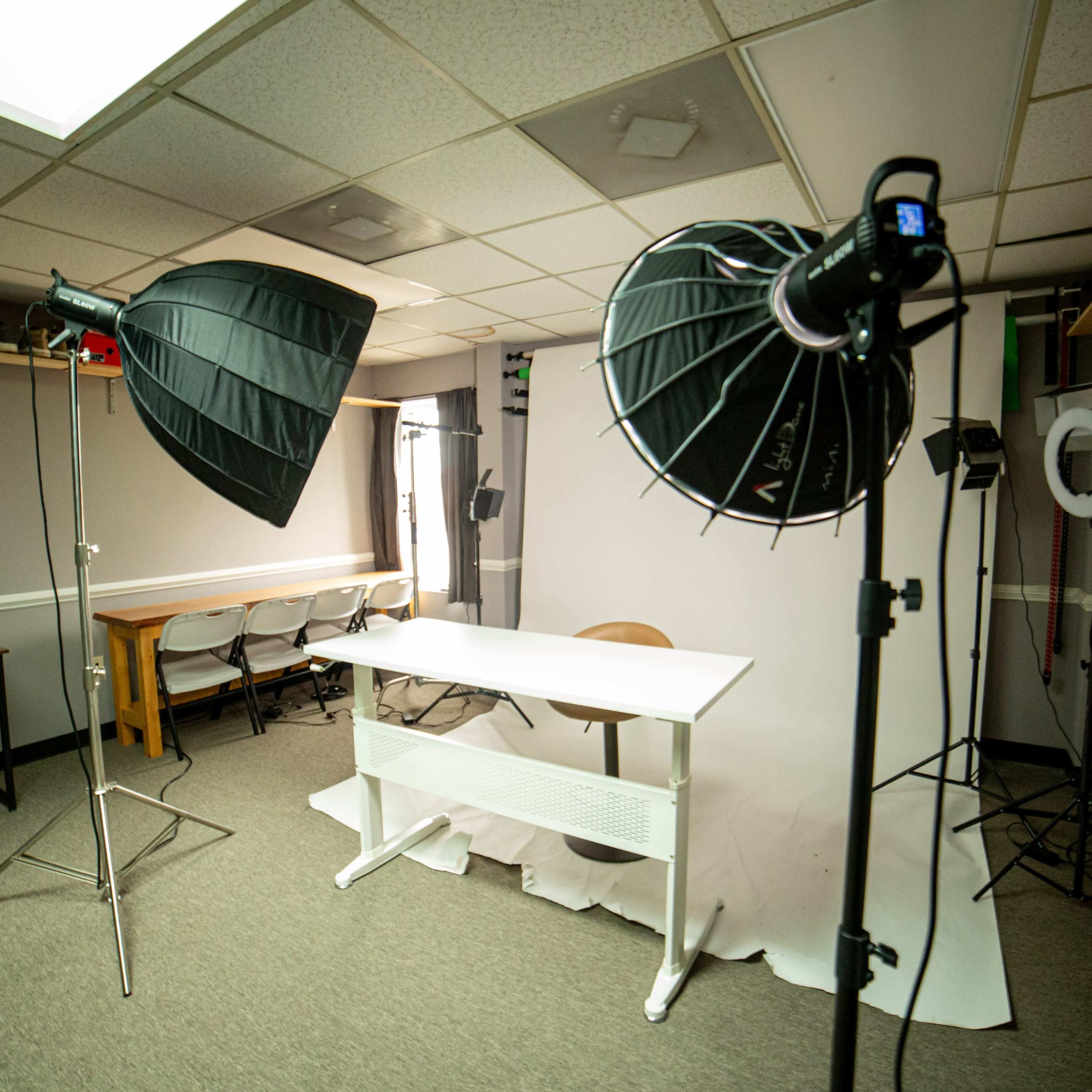 A photography studio setup features a white backdrop, a table with a chair, and multiple lighting equipment on either side.