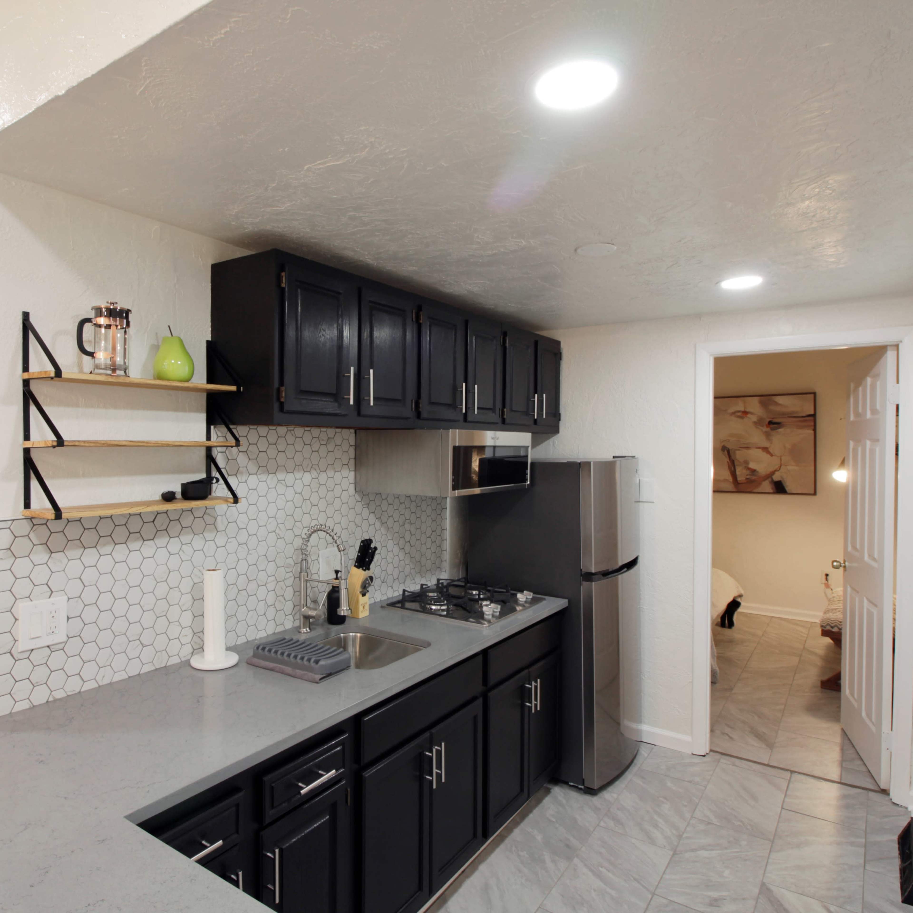 A modern kitchen featuring dark cabinetry, a gray countertop, and a hexagonal tile backsplash, with a doorway leading to another room in the background.