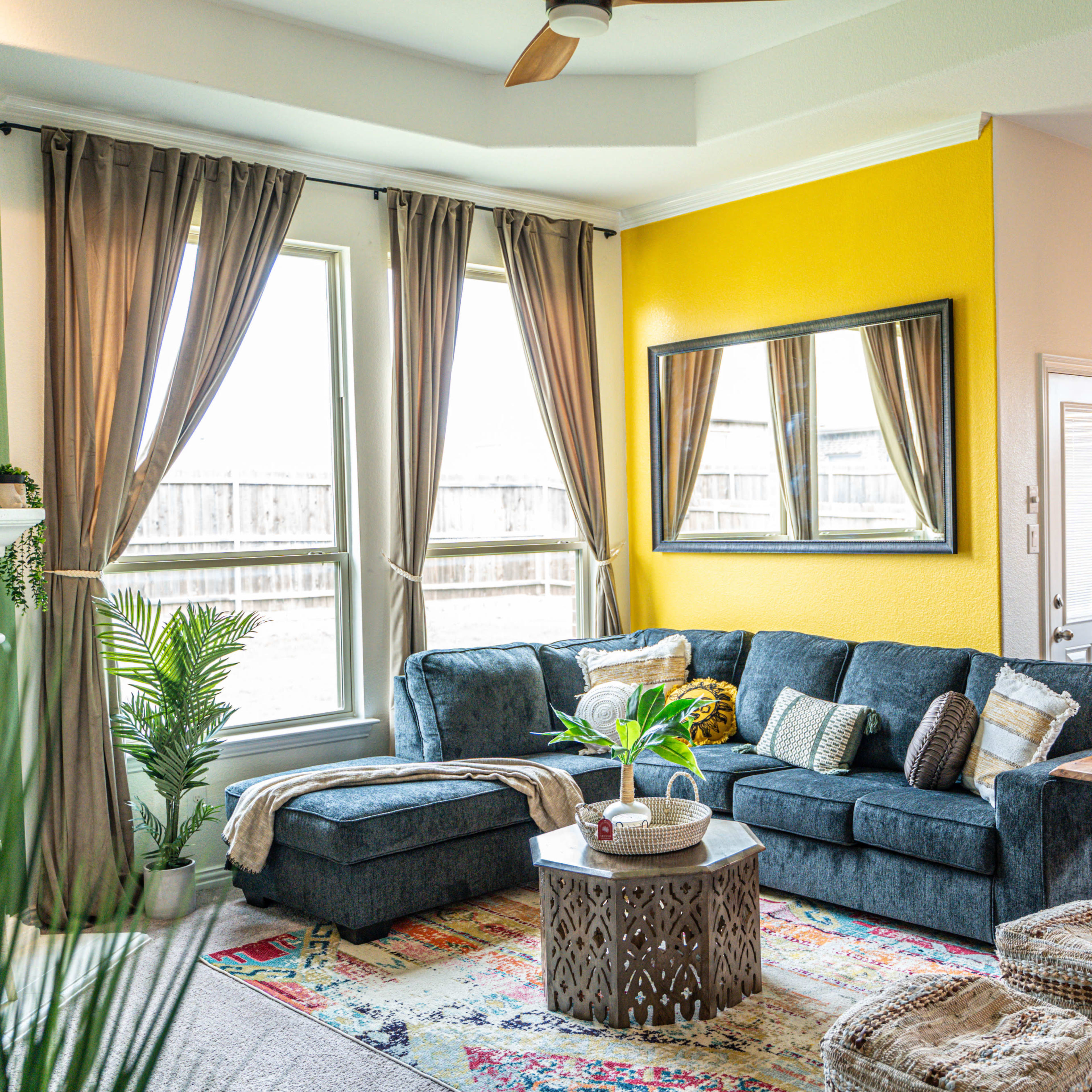 A modern living room features a sectional sofa, decorative pillows, a round coffee table, a large mirror, and a combination of natural light and vibrant wall colors.