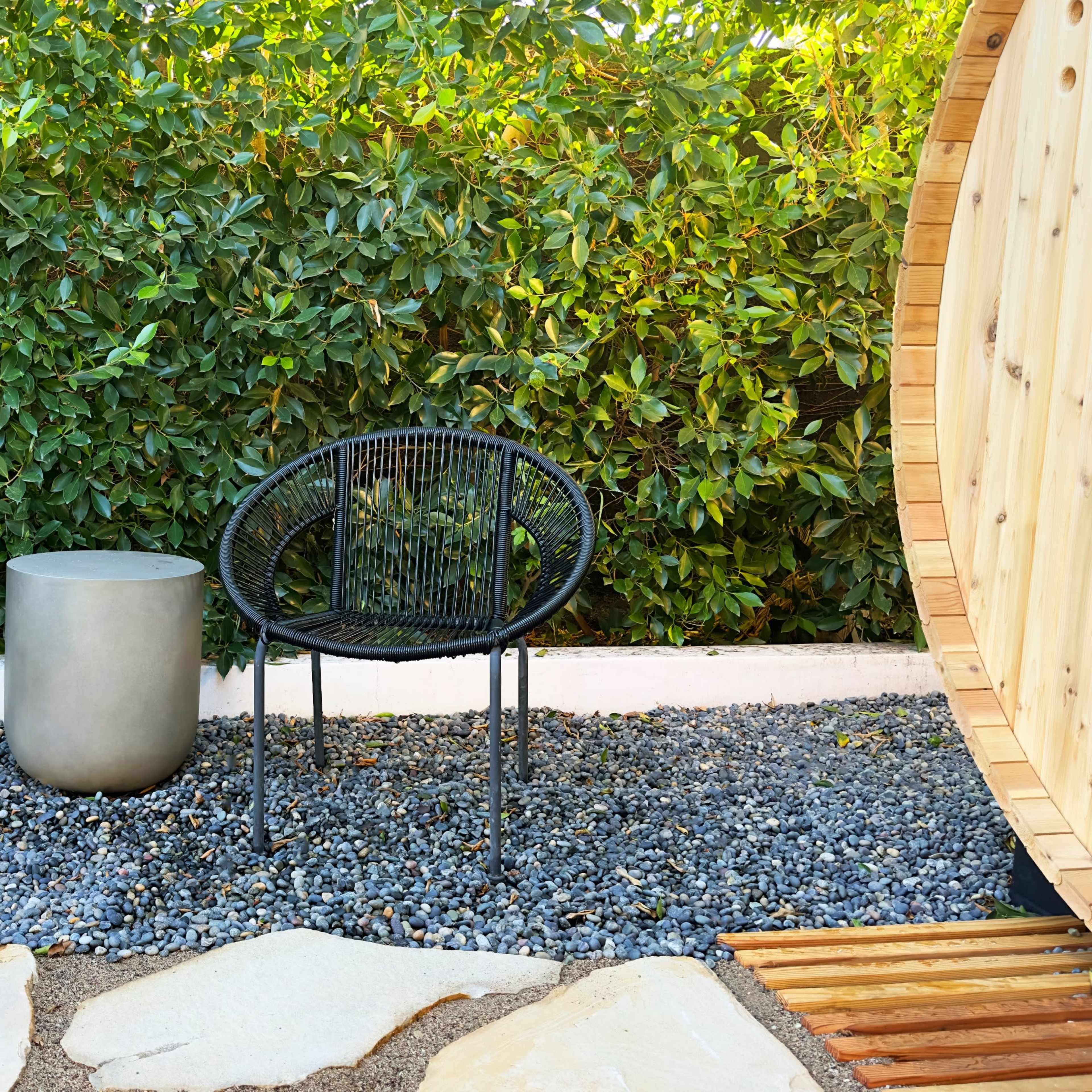Two black chairs and a round concrete table positioned on gravel near a wooden structure surrounded by lush green foliage.