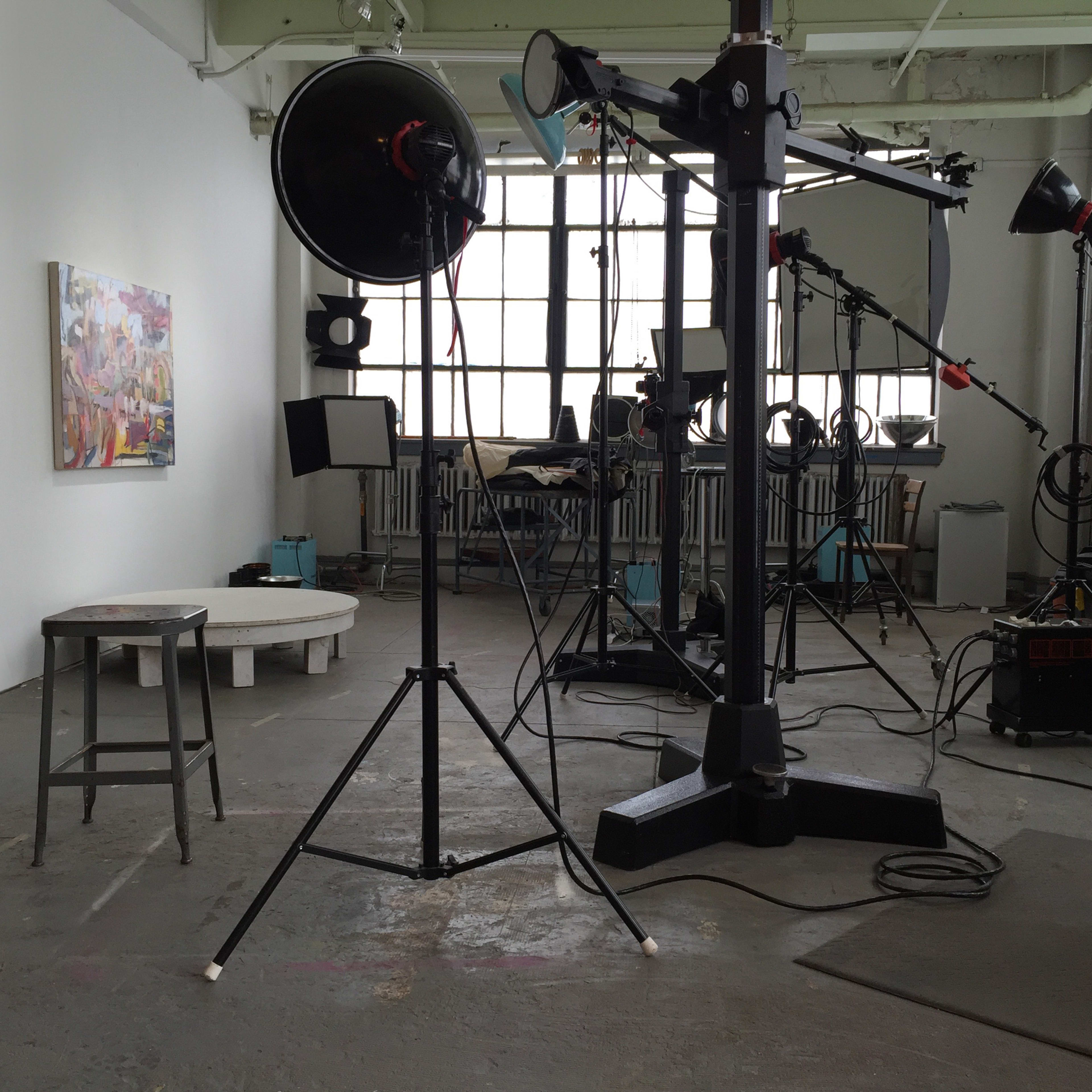 A photography studio with various lighting equipment, a stool, and a painting on the wall.