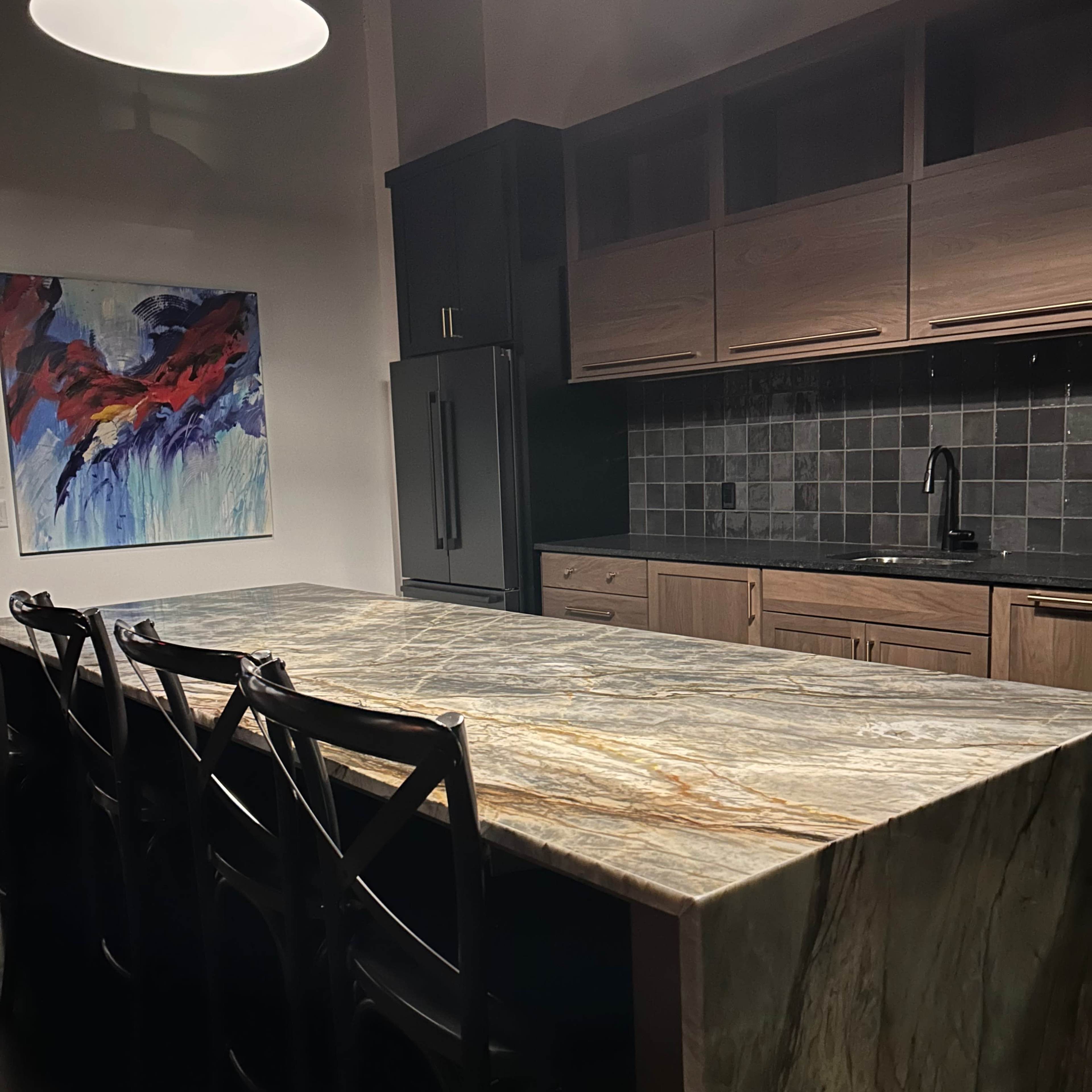 A large stone kitchen island with black chairs is positioned in a modern kitchen that features wooden cabinets and a colorful abstract painting on the wall.