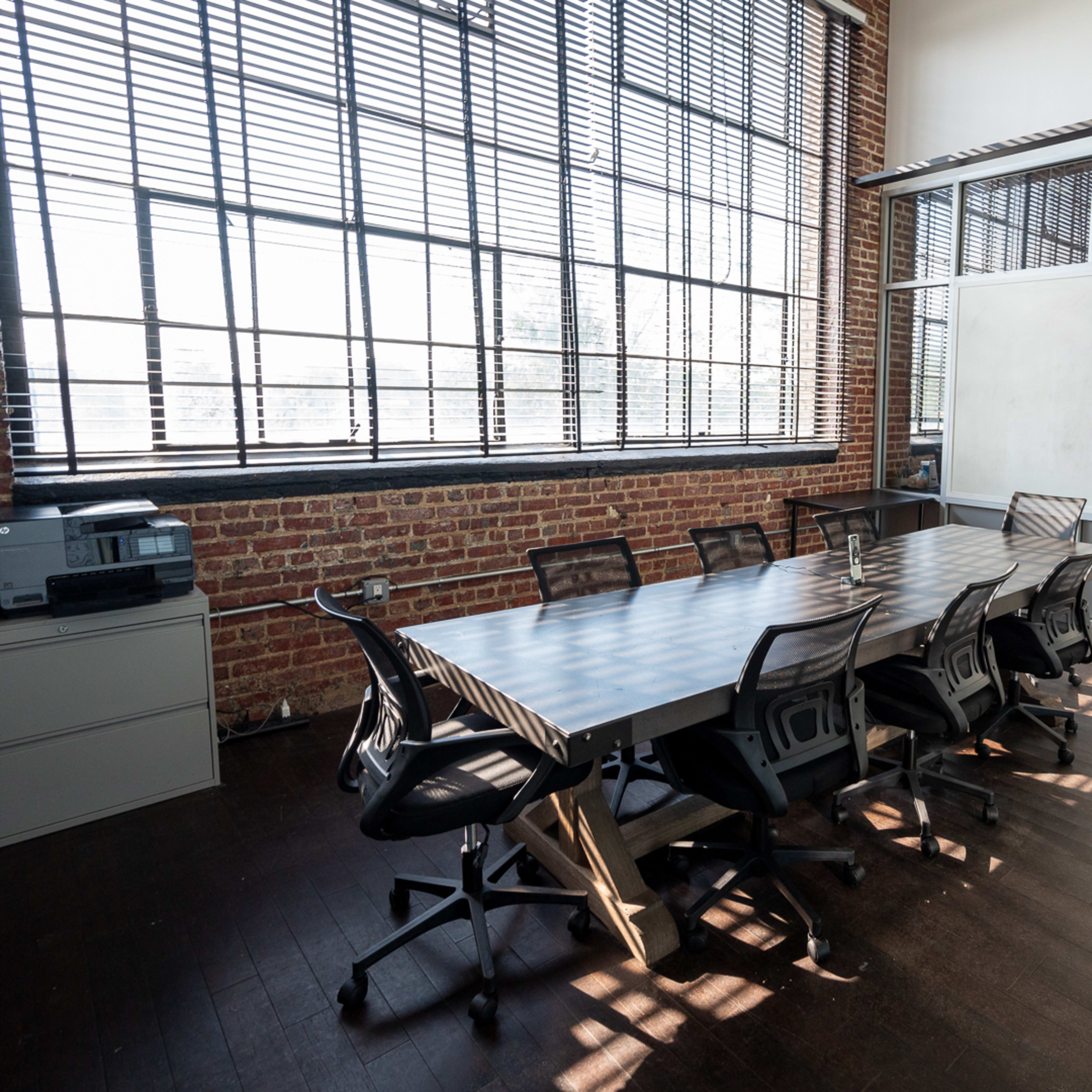 A conference room features a long table surrounded by chairs, with large windows and exposed brick walls.