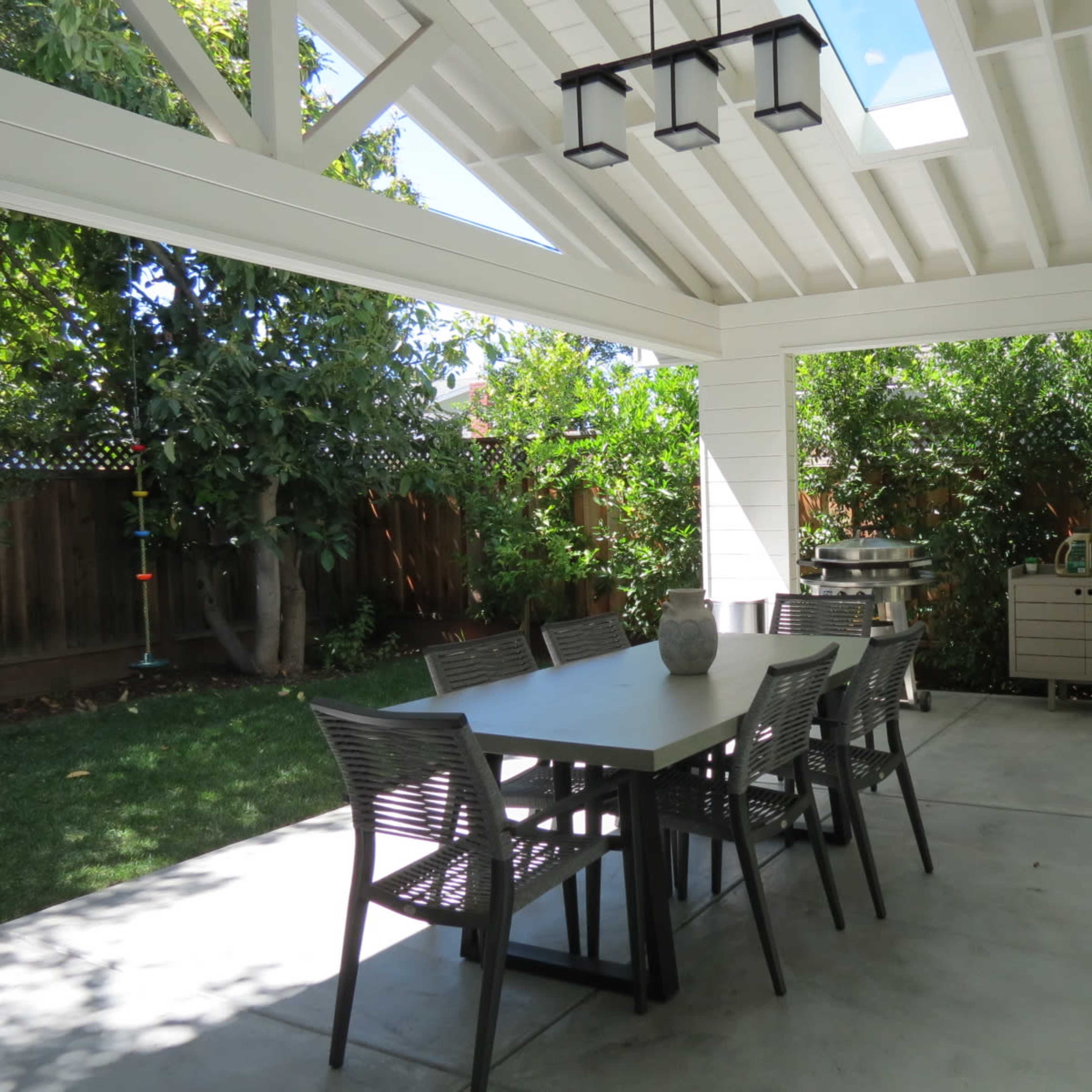 A modern outdoor dining area features a rectangular table surrounded by chairs under a covered patio with plants and greenery in the background.