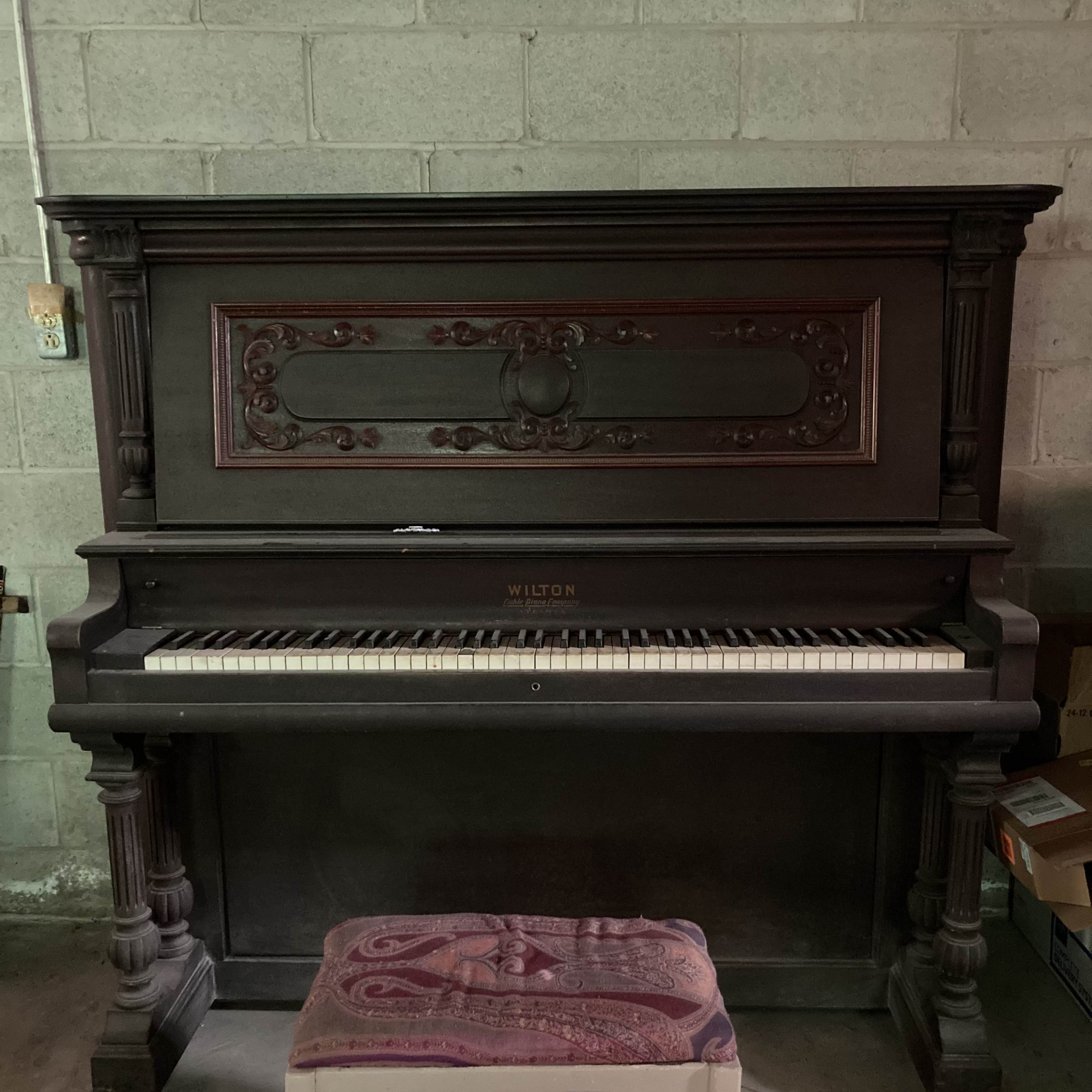 An antique Wilton piano with a decorative front is positioned against a concrete wall, accompanied by a patterned stool in front.