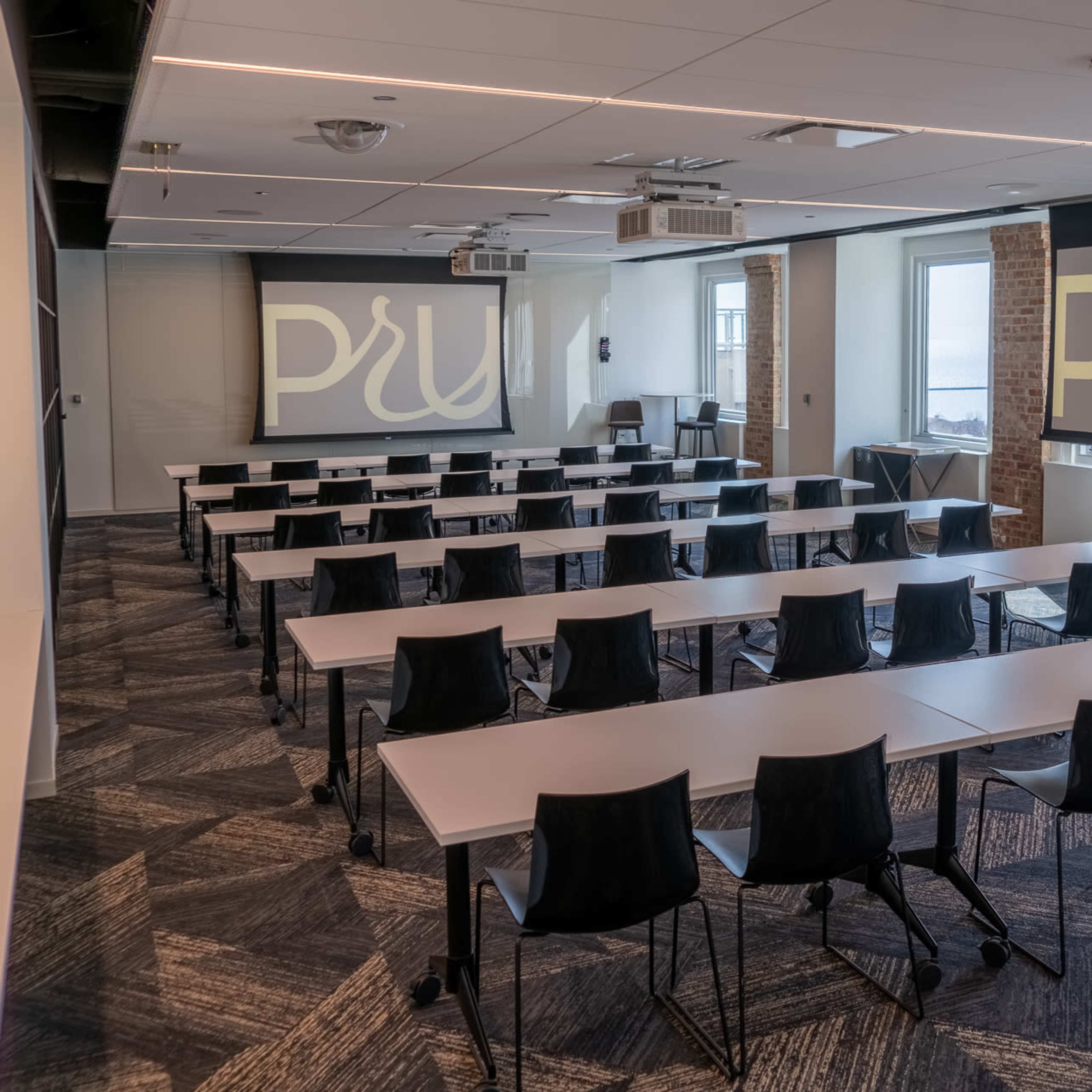 A conference room set up with rows of tables and chairs, featuring two large projection screens at the front displaying the letters "PRU."