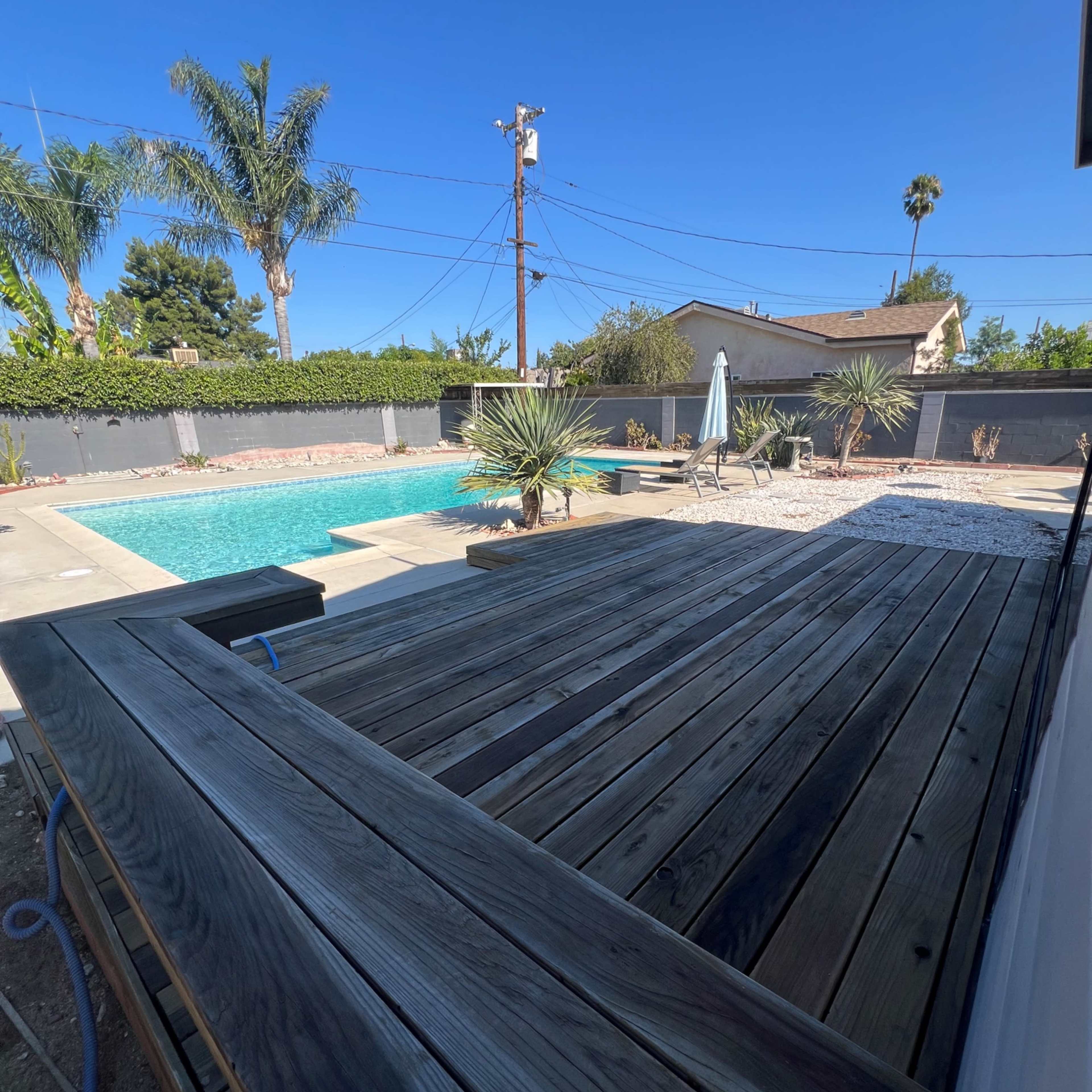 A wooden deck overlooks a swimming pool surrounded by palm trees and desert plants in a residential area.