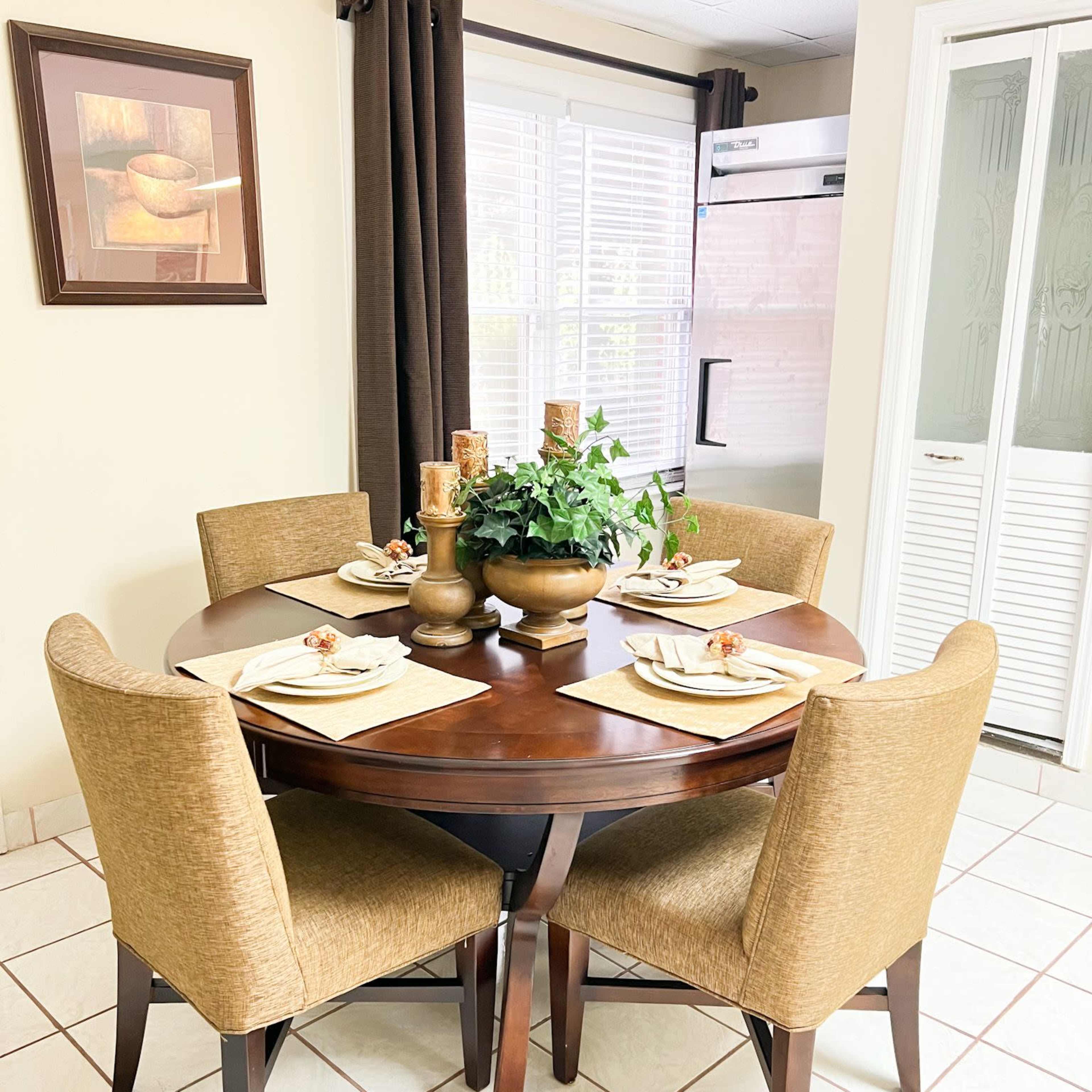A round wooden dining table is set for four, surrounded by beige upholstered chairs, with a plant centerpiece and curtains framing a window in a well-lit kitchen.