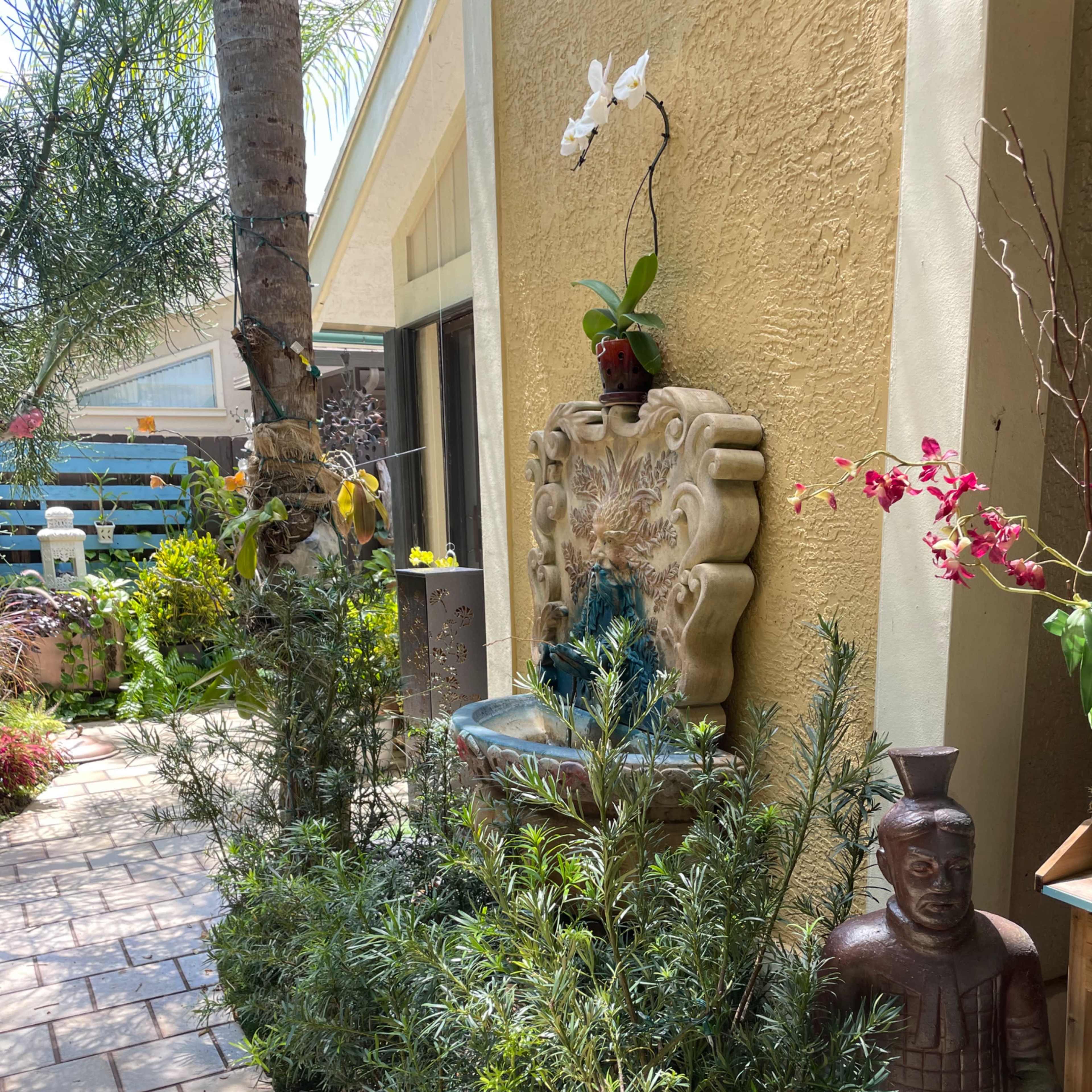 A decorative fountain is positioned against a yellow wall, surrounded by lush greenery and a stone figure, with a palm tree observable nearby.