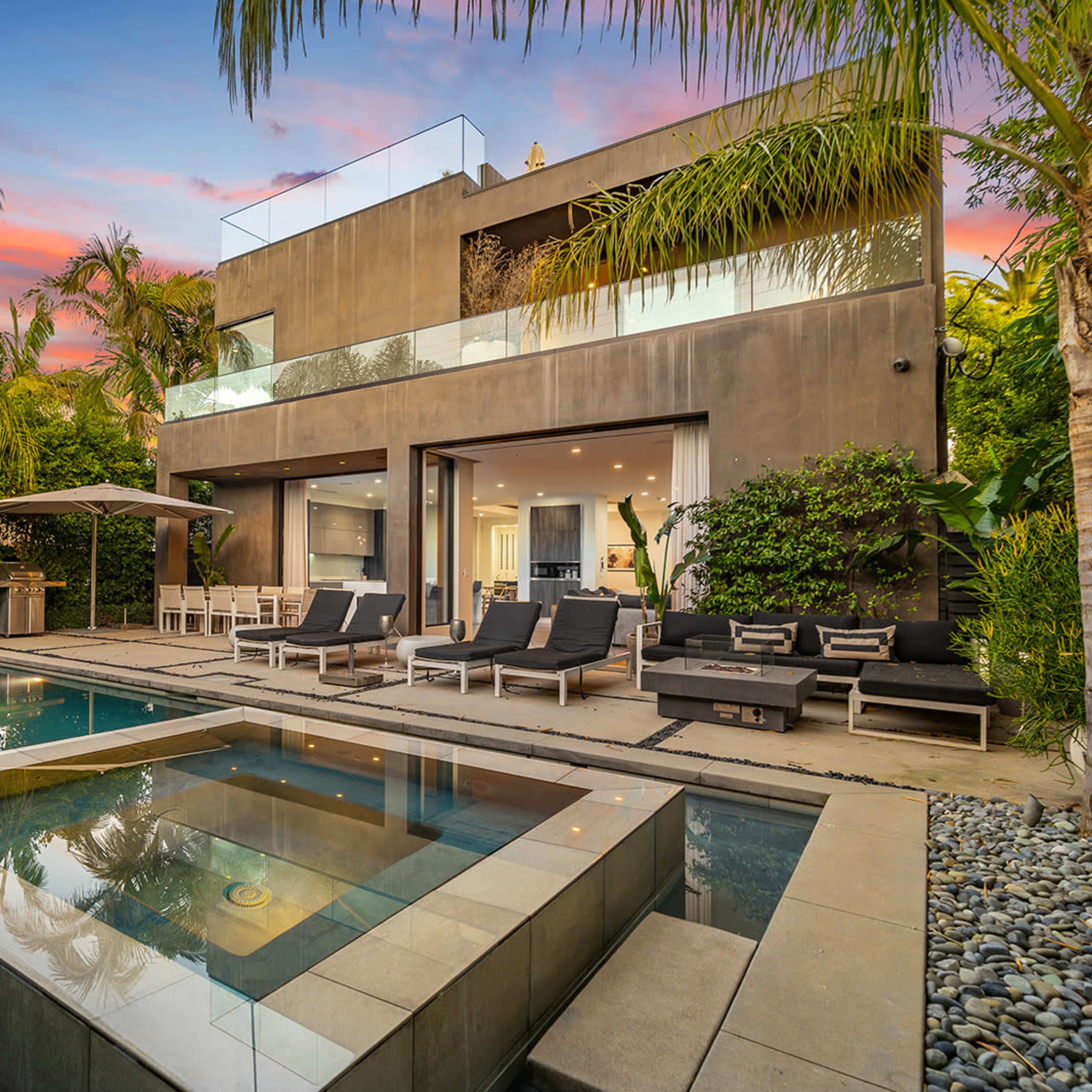 A modern home with a swimming pool and spa area, surrounded by palm trees and lush greenery, under a colorful sunset sky.