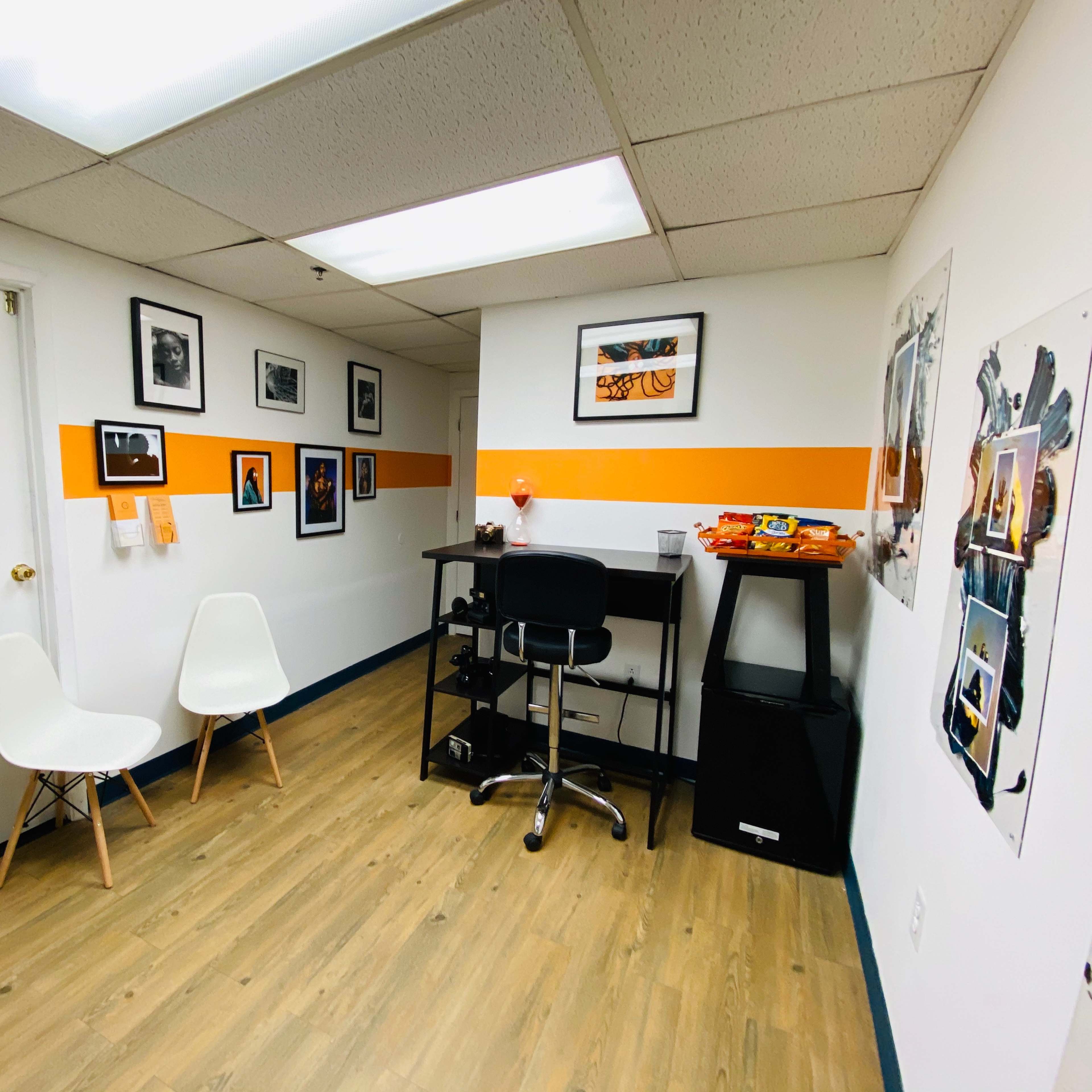 A small office space with two white chairs, a black desk and chair, and framed photos on the walls, featuring an orange accent stripe.