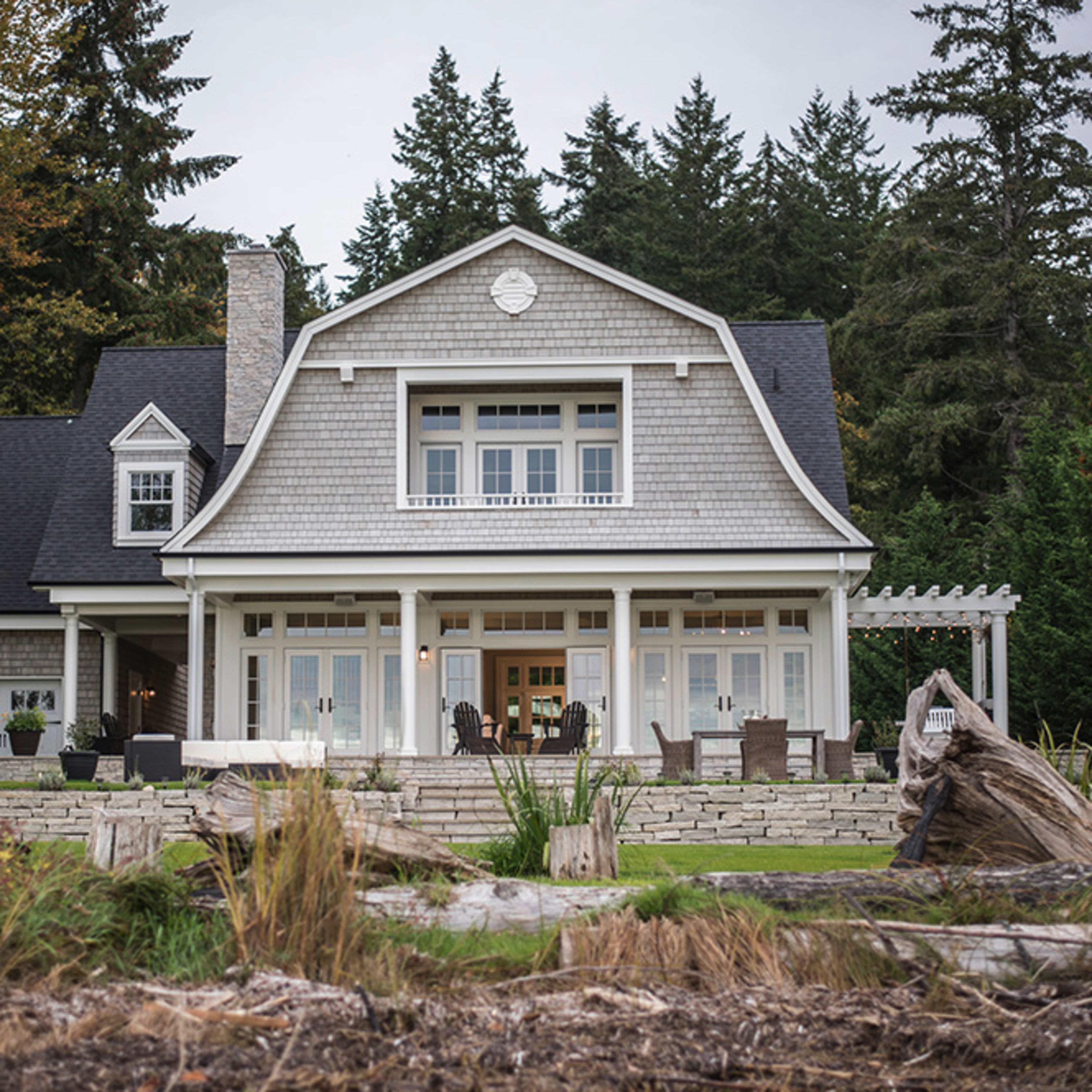 A coastal-style house with a gray shingle exterior features a spacious front porch and is surrounded by trees and landscaping.