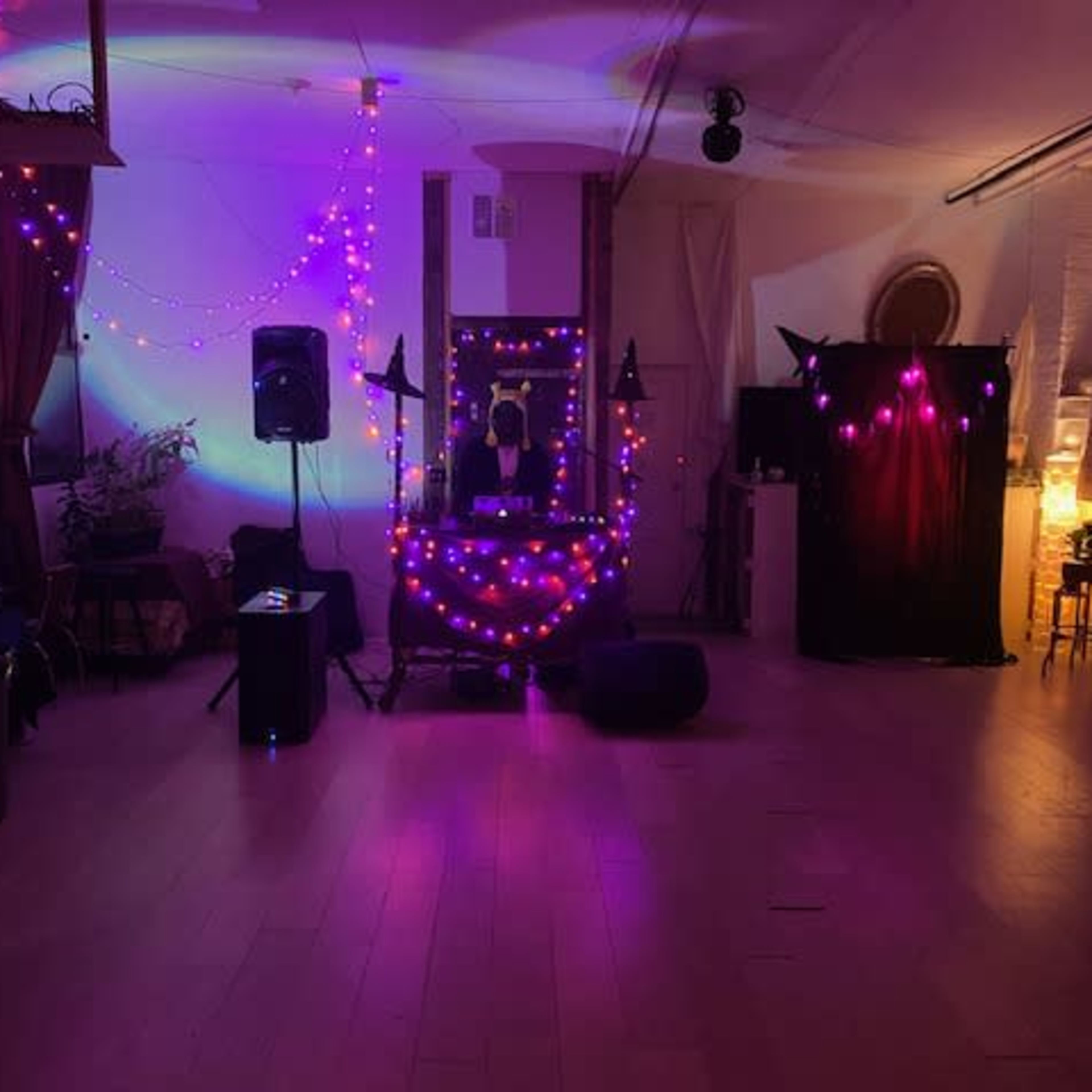 A dimly lit room decorated with colorful string lights, featuring a DJ setup with speakers and a screen draped in black.