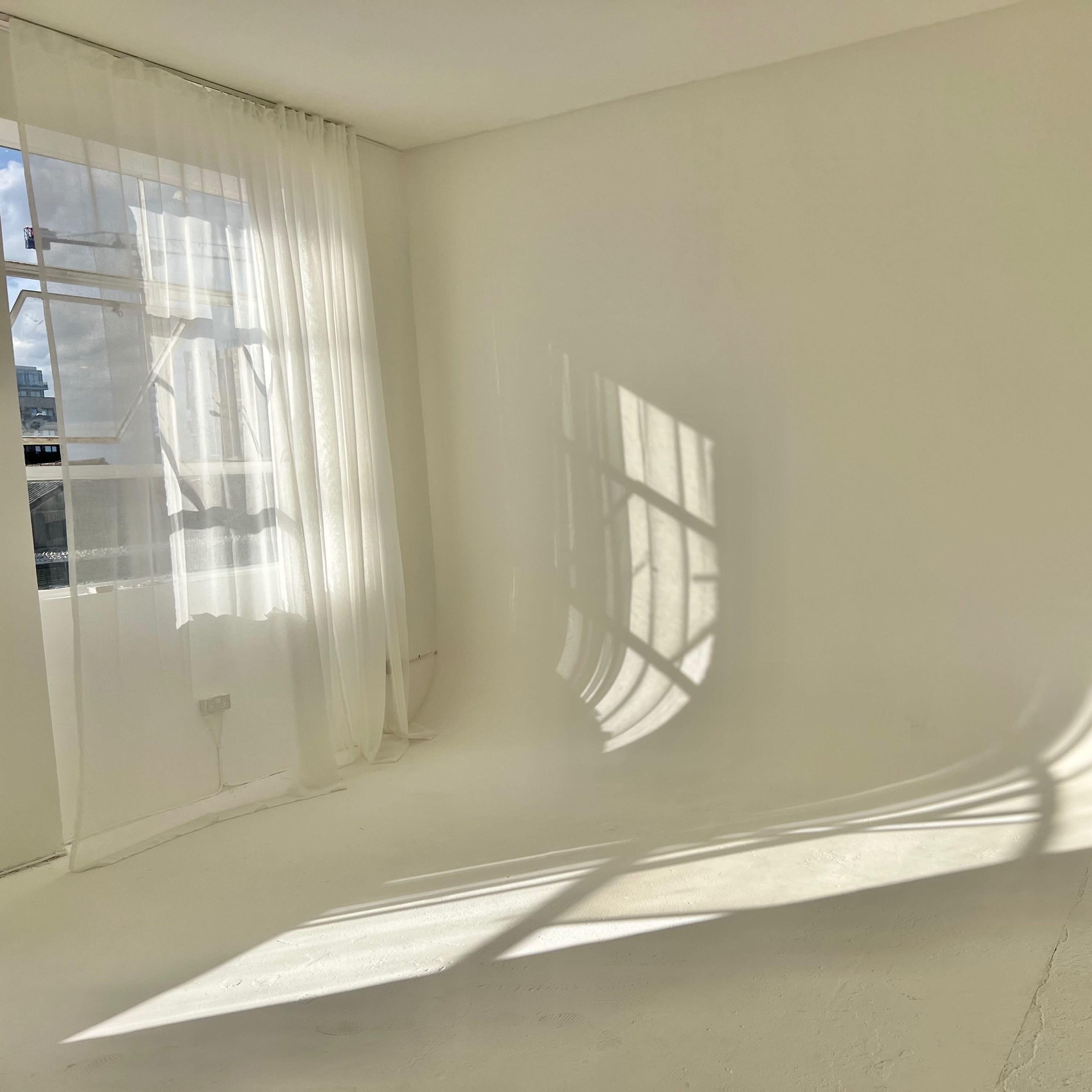 A spacious, bright, white room with a large window allowing sunlight to create shadows on the floor, and a beige couch positioned against one wall.