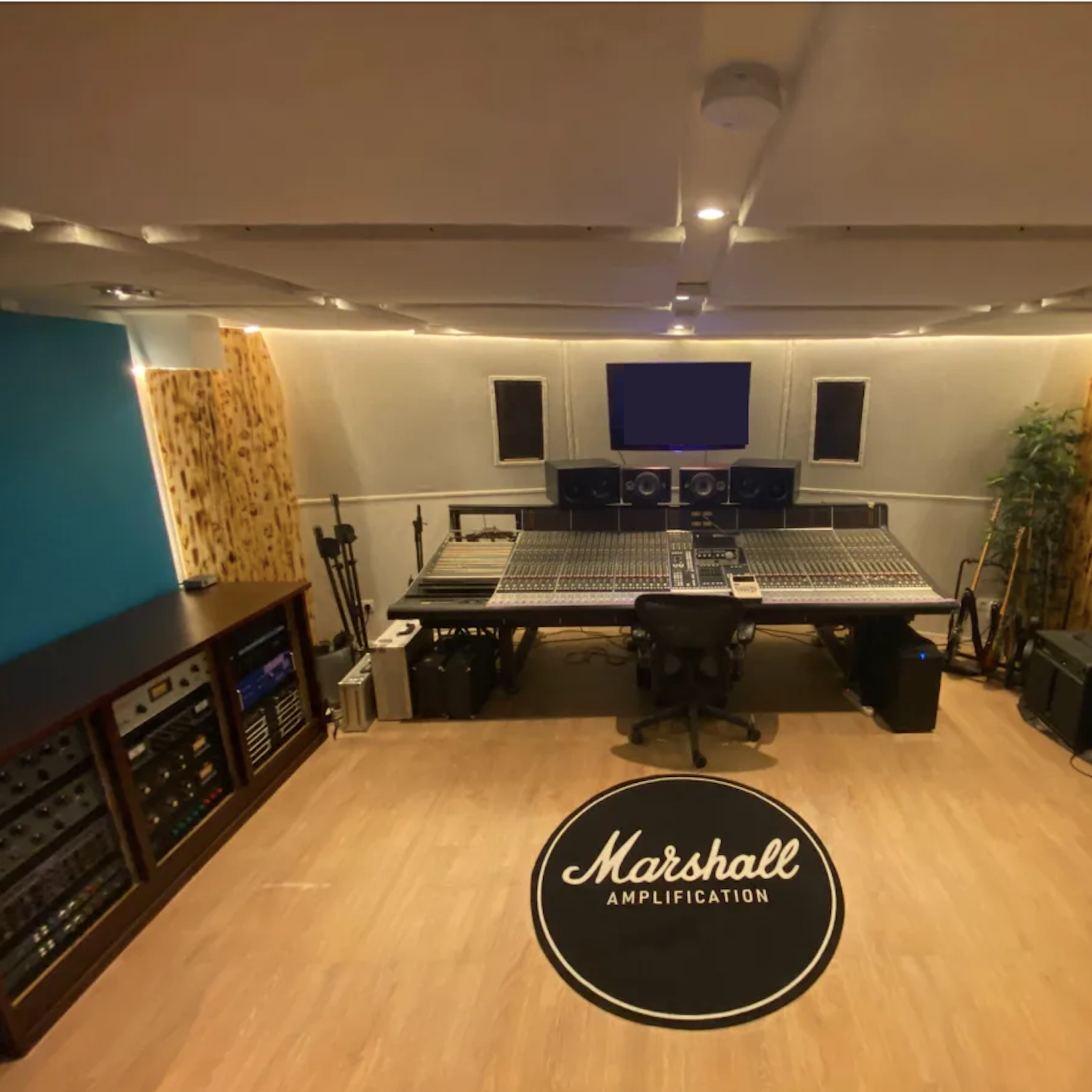 A music recording studio with a central mixing console, surrounded by audio equipment and a large logo on the floor.
