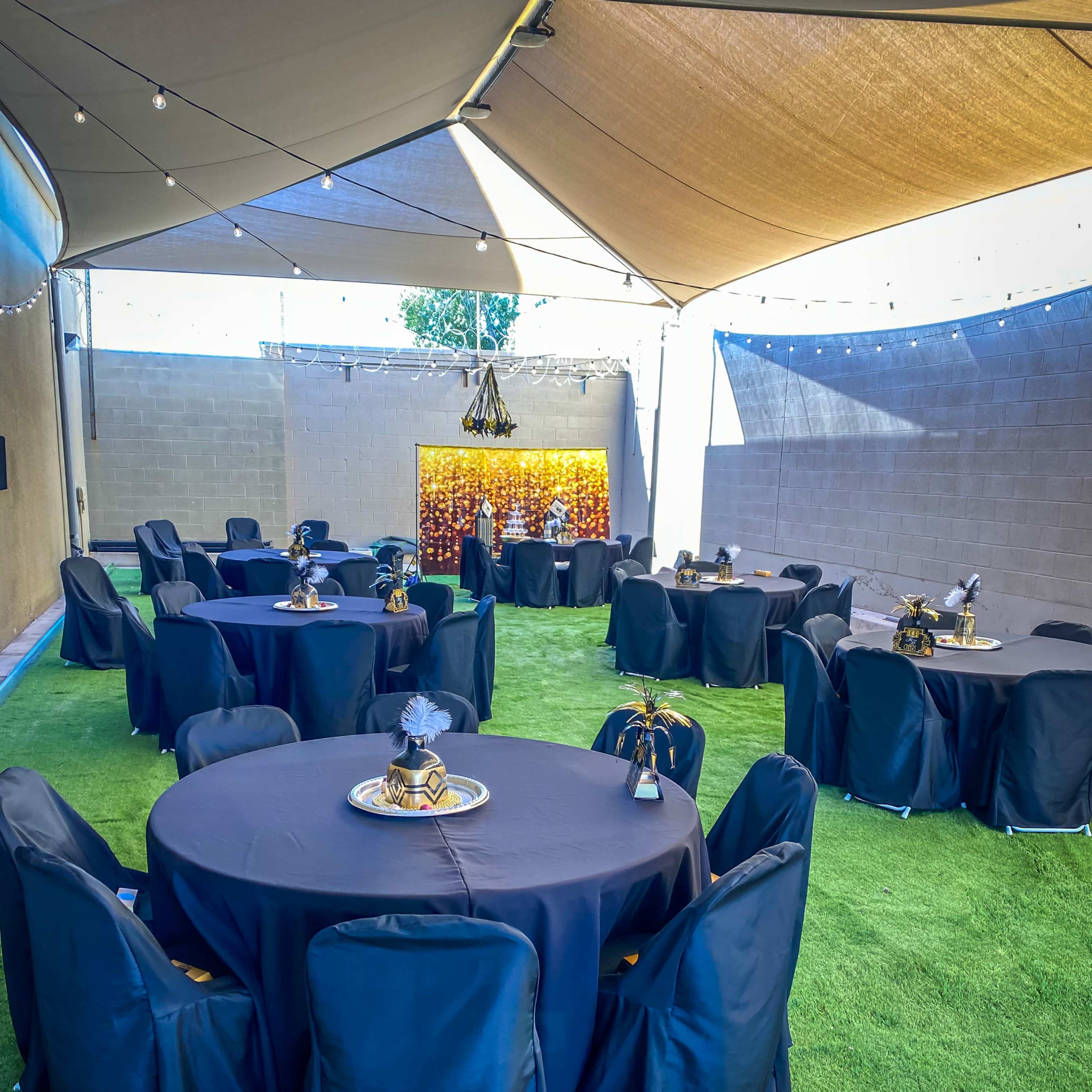 A patio space with several round tables covered in black tablecloths, surrounded by black chairs, and decorated with centerpieces, all beneath a canopy and string lights.