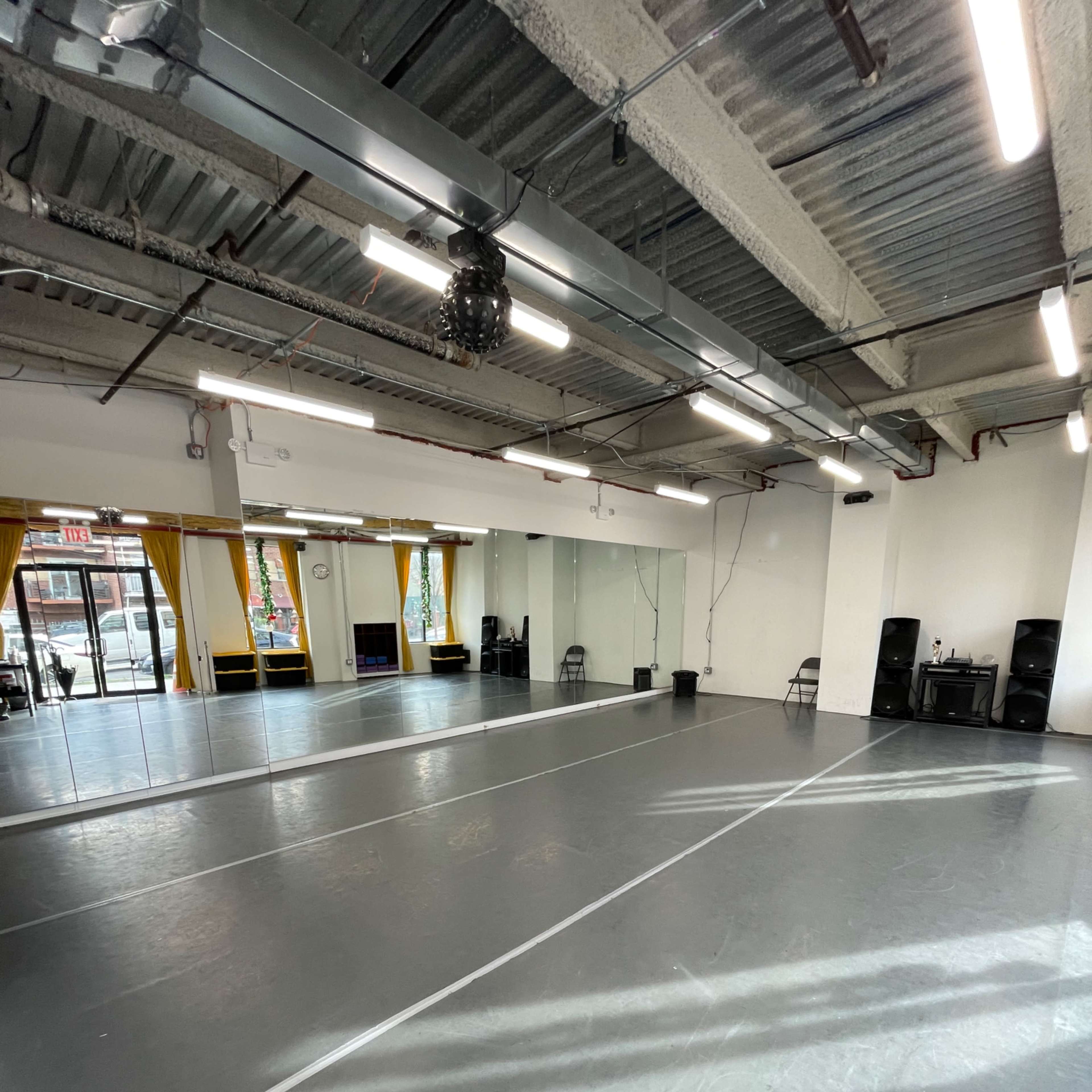 A spacious dance studio with gray flooring, wall mirrors, and large windows allowing natural light to enter.