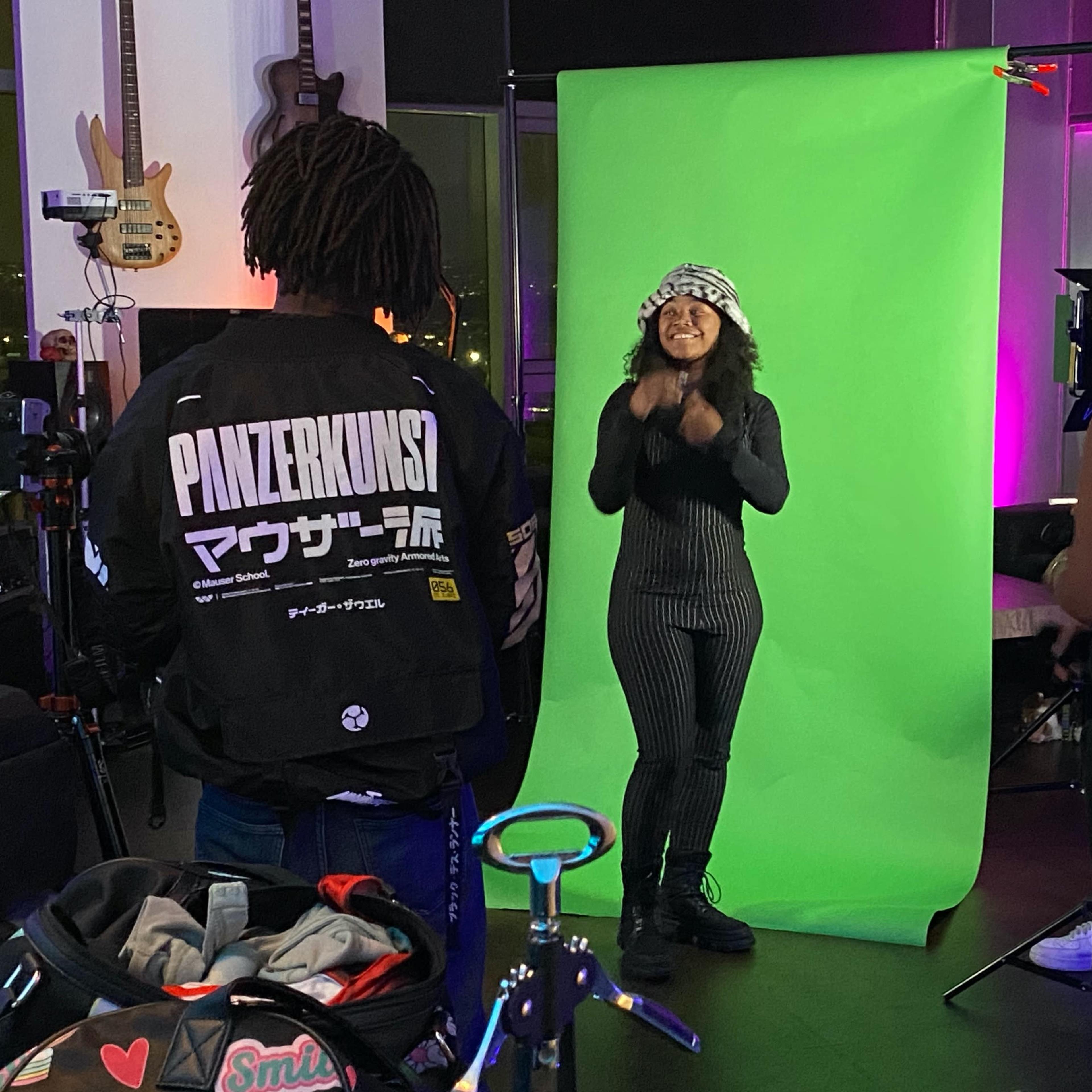 A group of people is in a studio setting, with one person in front of a green backdrop while two others engage with her, and a fourth person sits nearby.