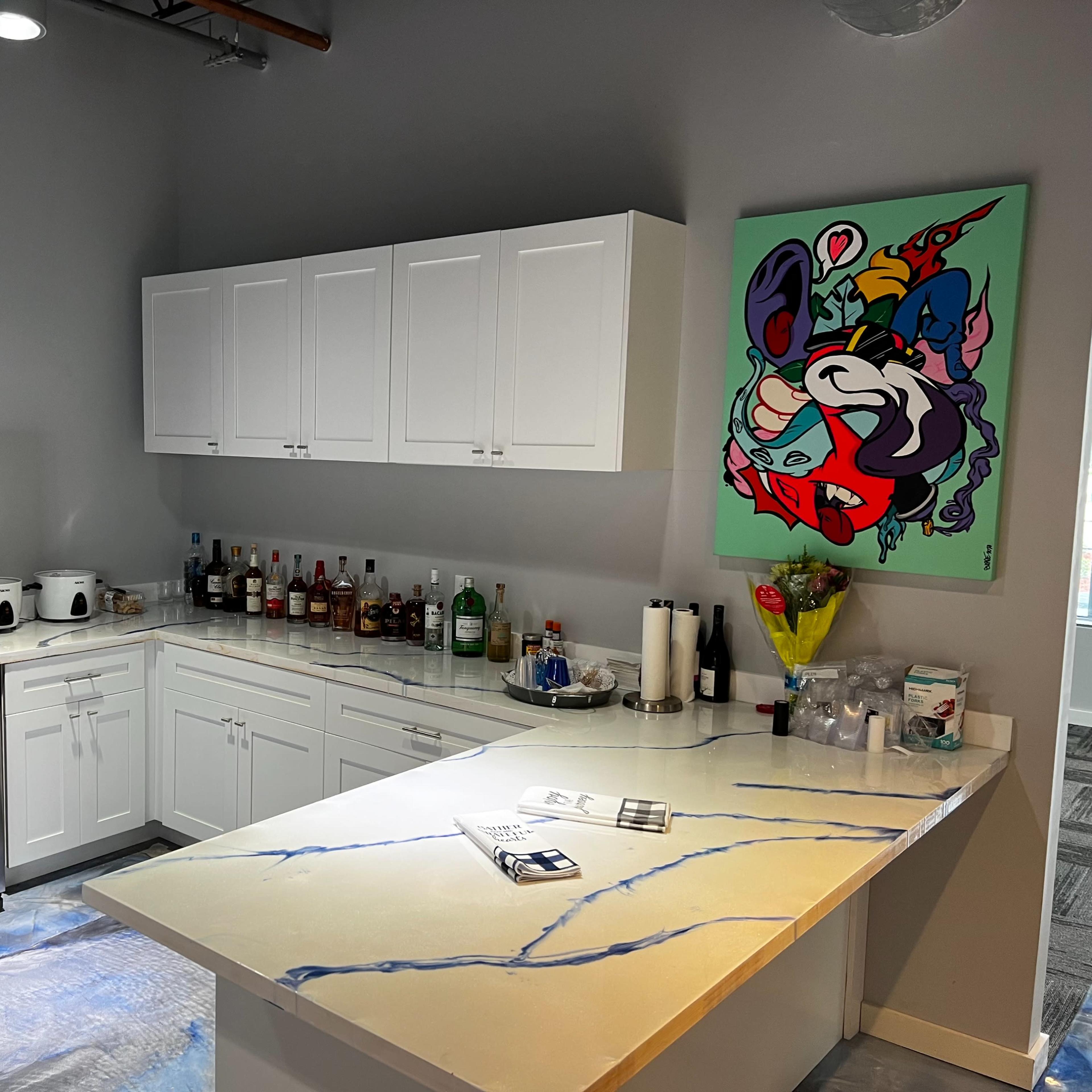A modern kitchen features a stainless steel refrigerator, white cabinetry, a marble countertop with blue veining, and a colorful abstract painting on the wall.