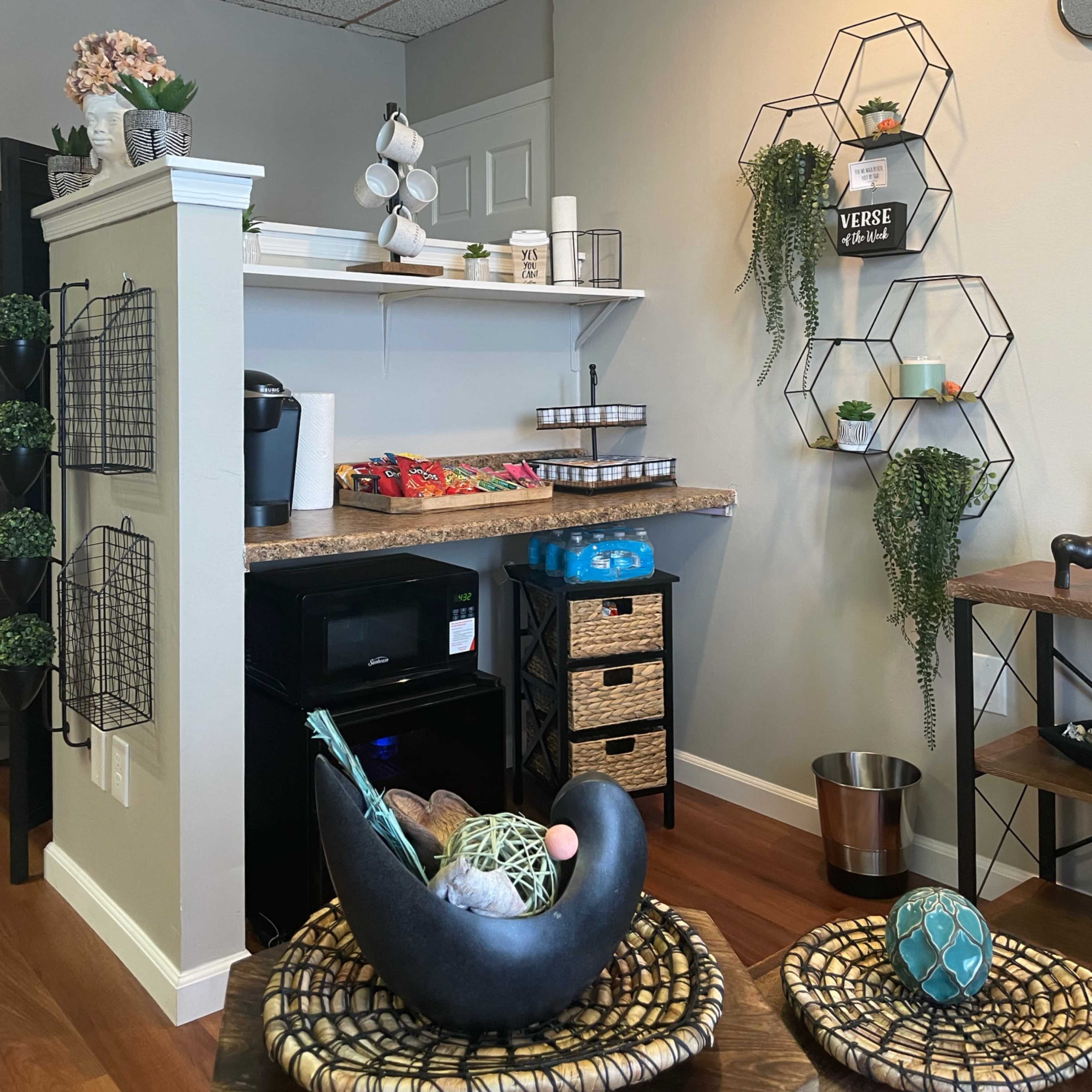 A small kitchen area with a countertop displaying snacks, a coffee machine, and decorative wall shelves featuring plants and a framed verse.