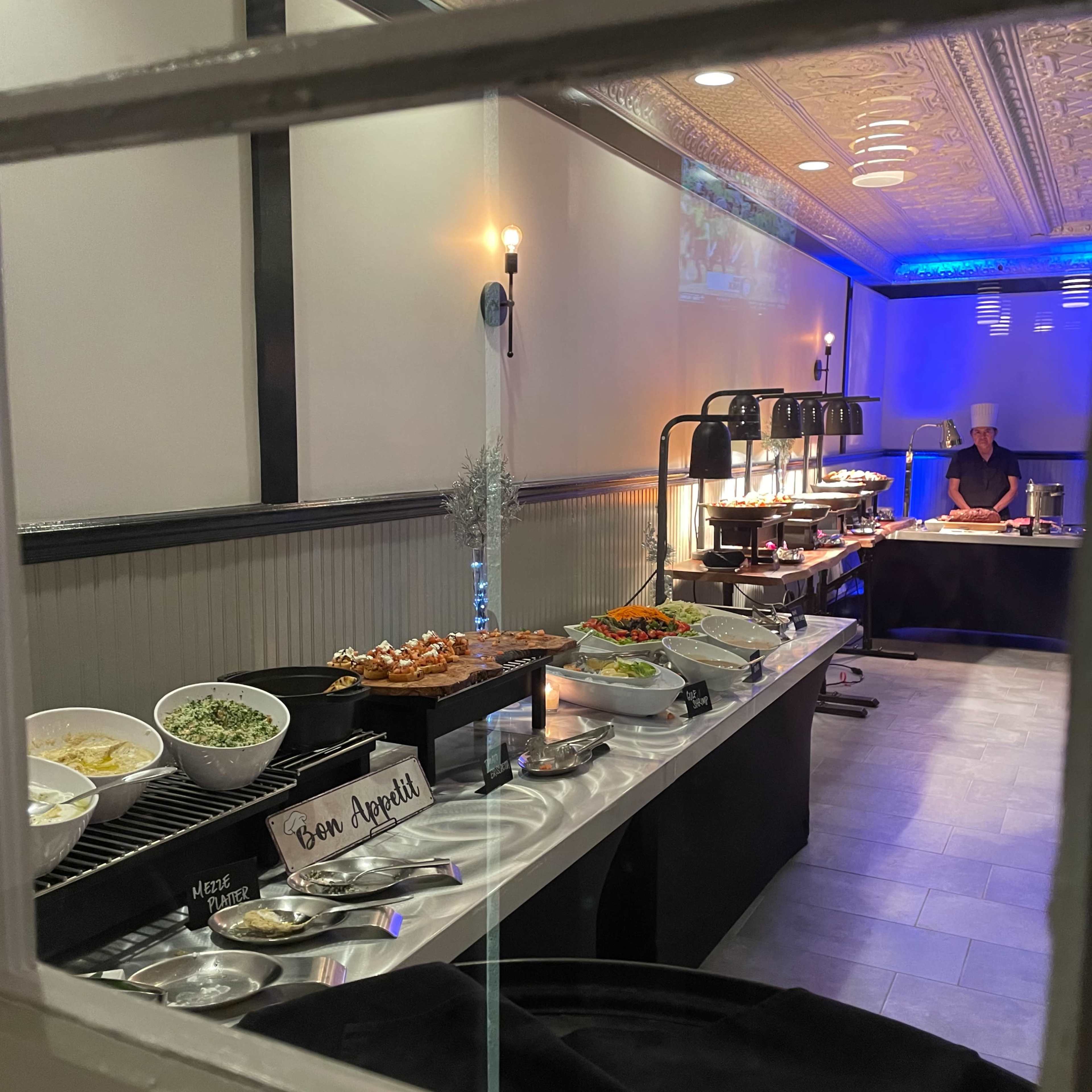 A buffet setup is visible through a window, featuring various dishes on a black tablecloth and a chef preparing food in the background.