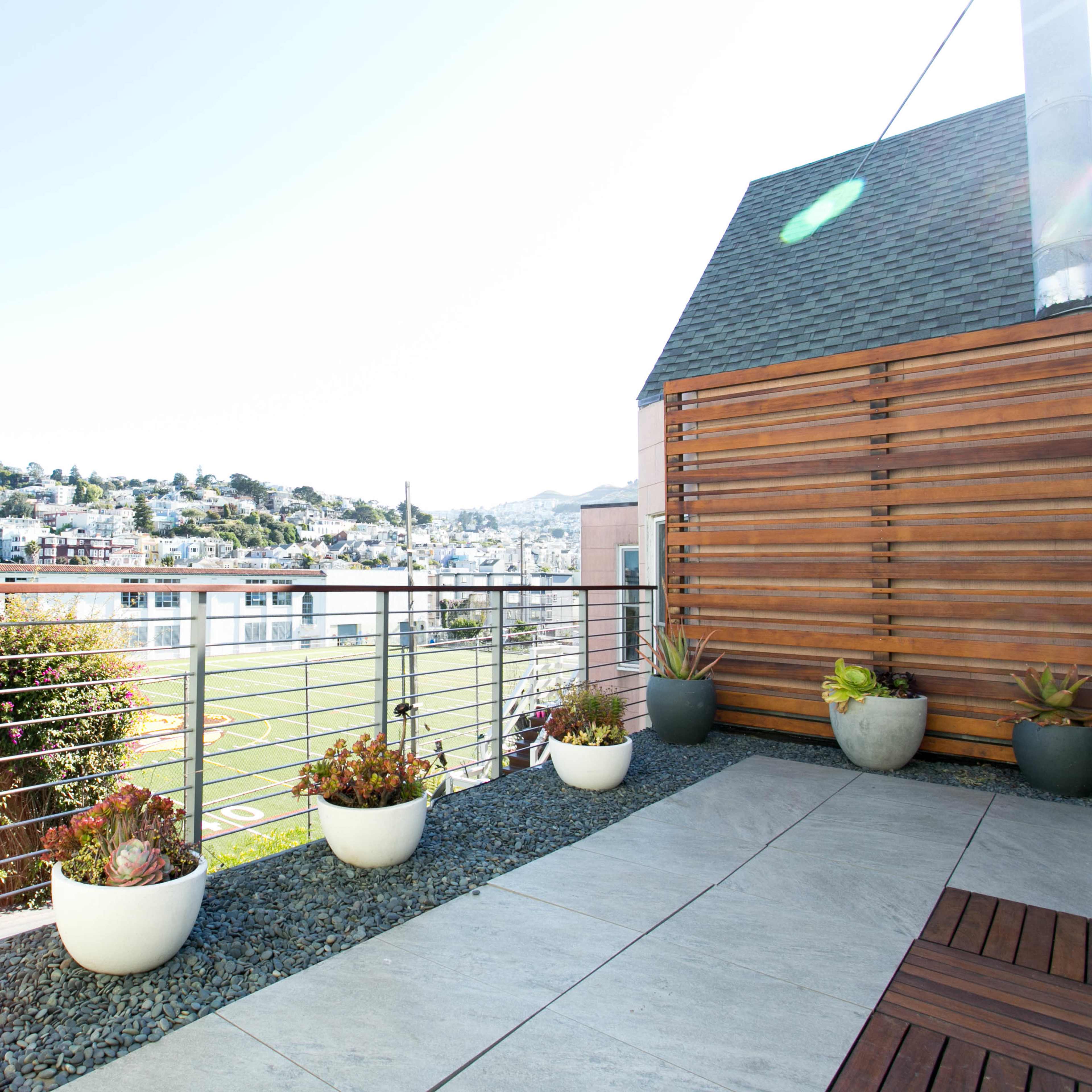 A rooftop terrace features potted plants along a railing, with a view of a neighborhood and a sports field in the background.