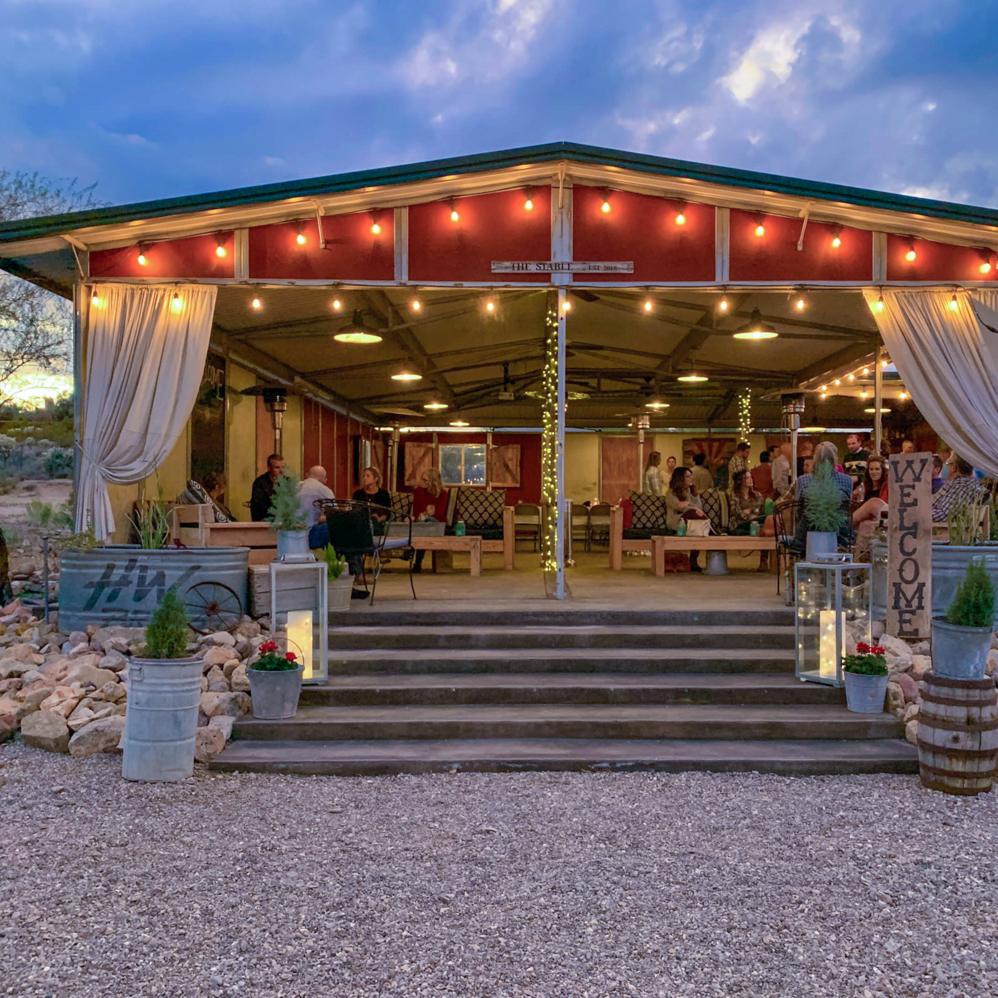 A rustic outdoor venue with a covered patio, adorned with string lights and hosting a gathering of people, is surrounded by desert scenery.