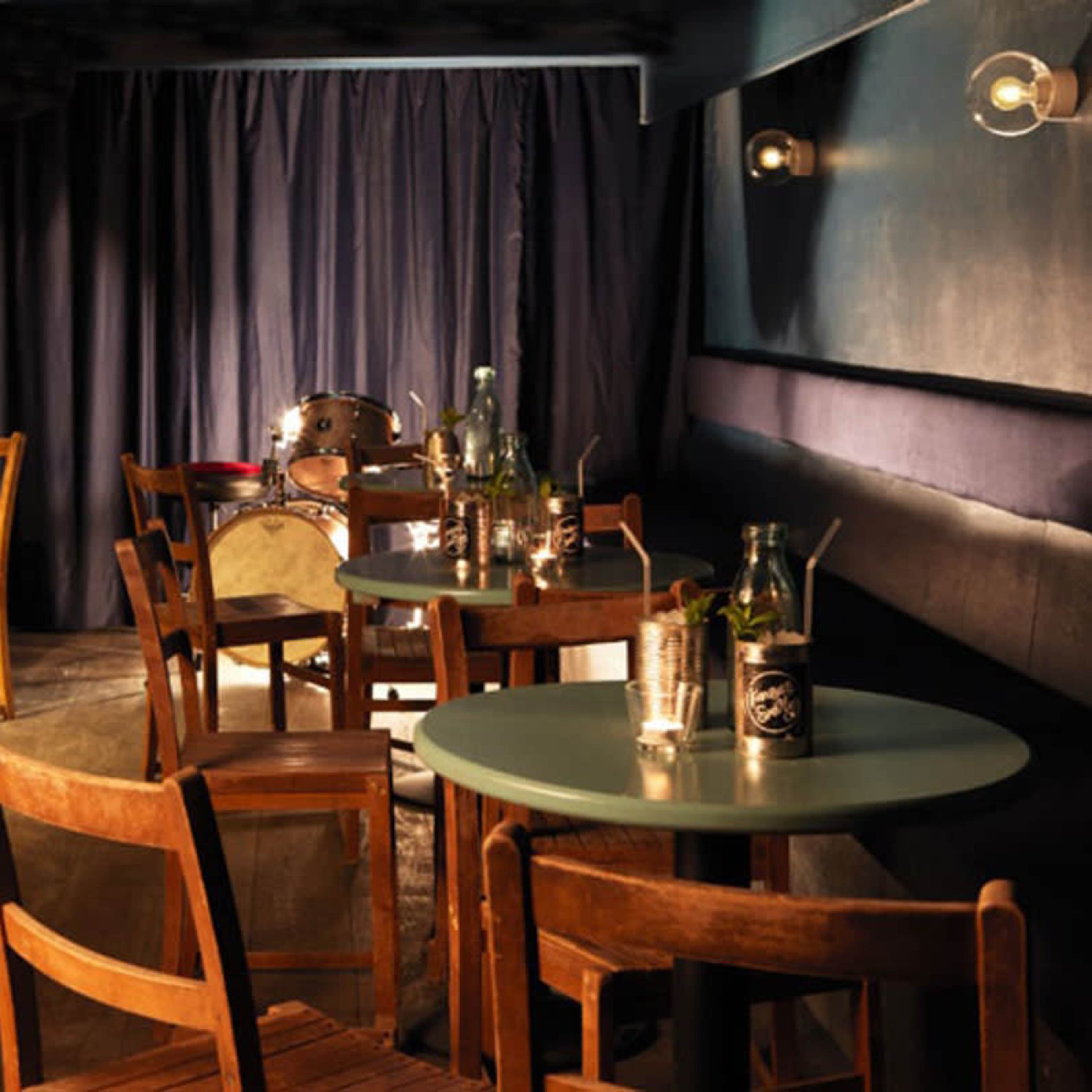 A small, dimly lit bar area features wooden tables and chairs, with a drum set visible in the background.