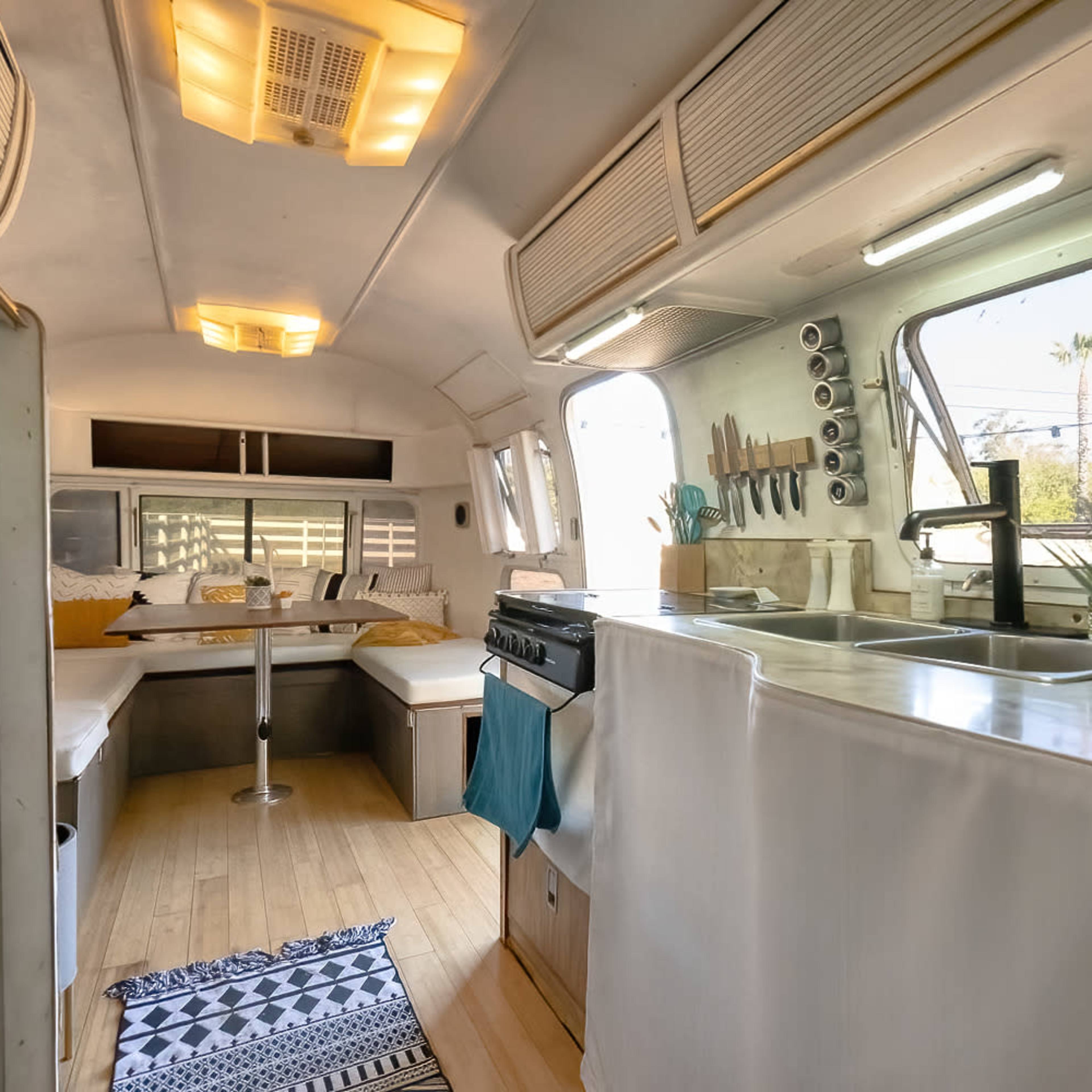The interior of a vintage trailer features a dining area with a table, kitchenette with a stove and sink, and large windows offering natural light.