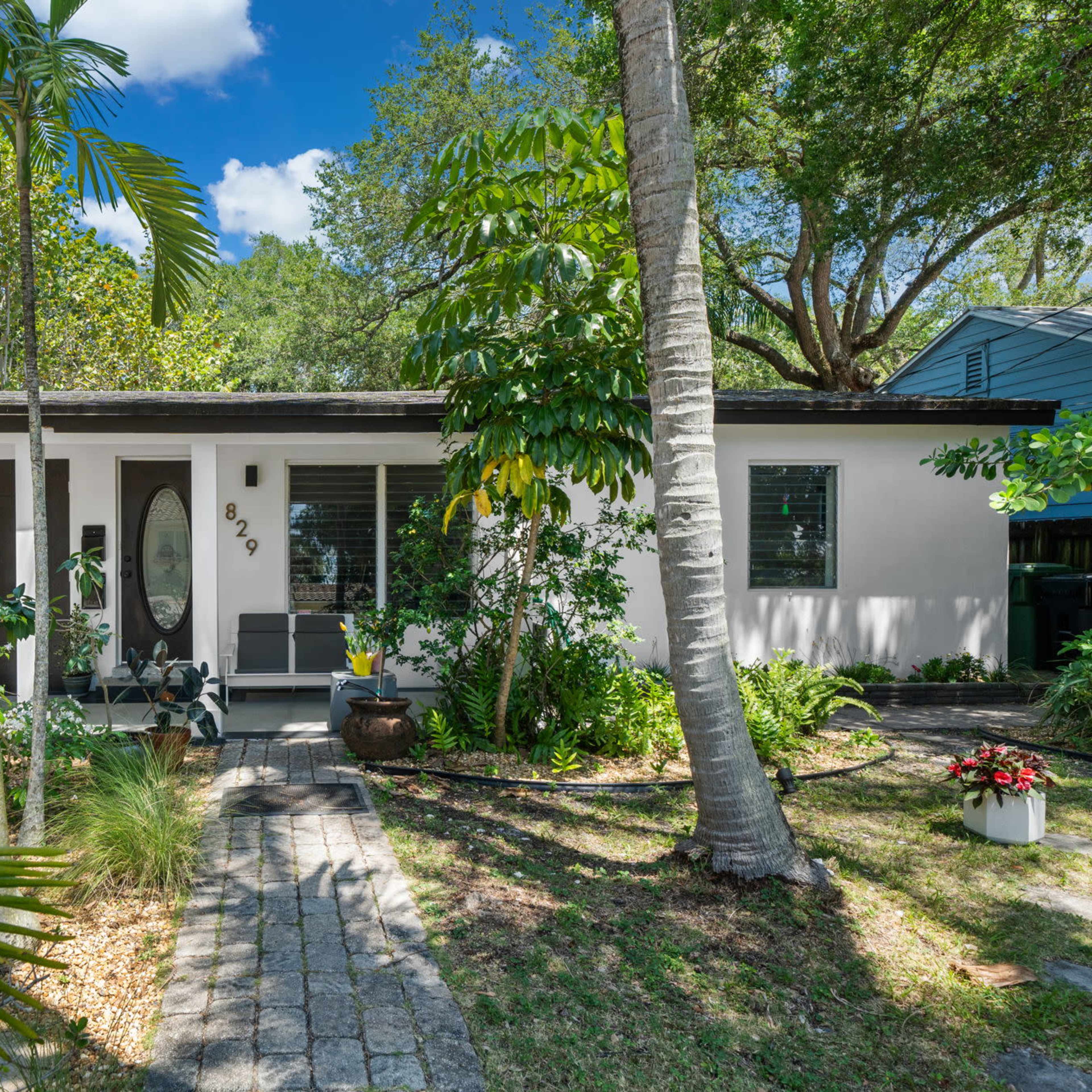 A single-story house with a front porch, surrounded by greenery and palm trees.