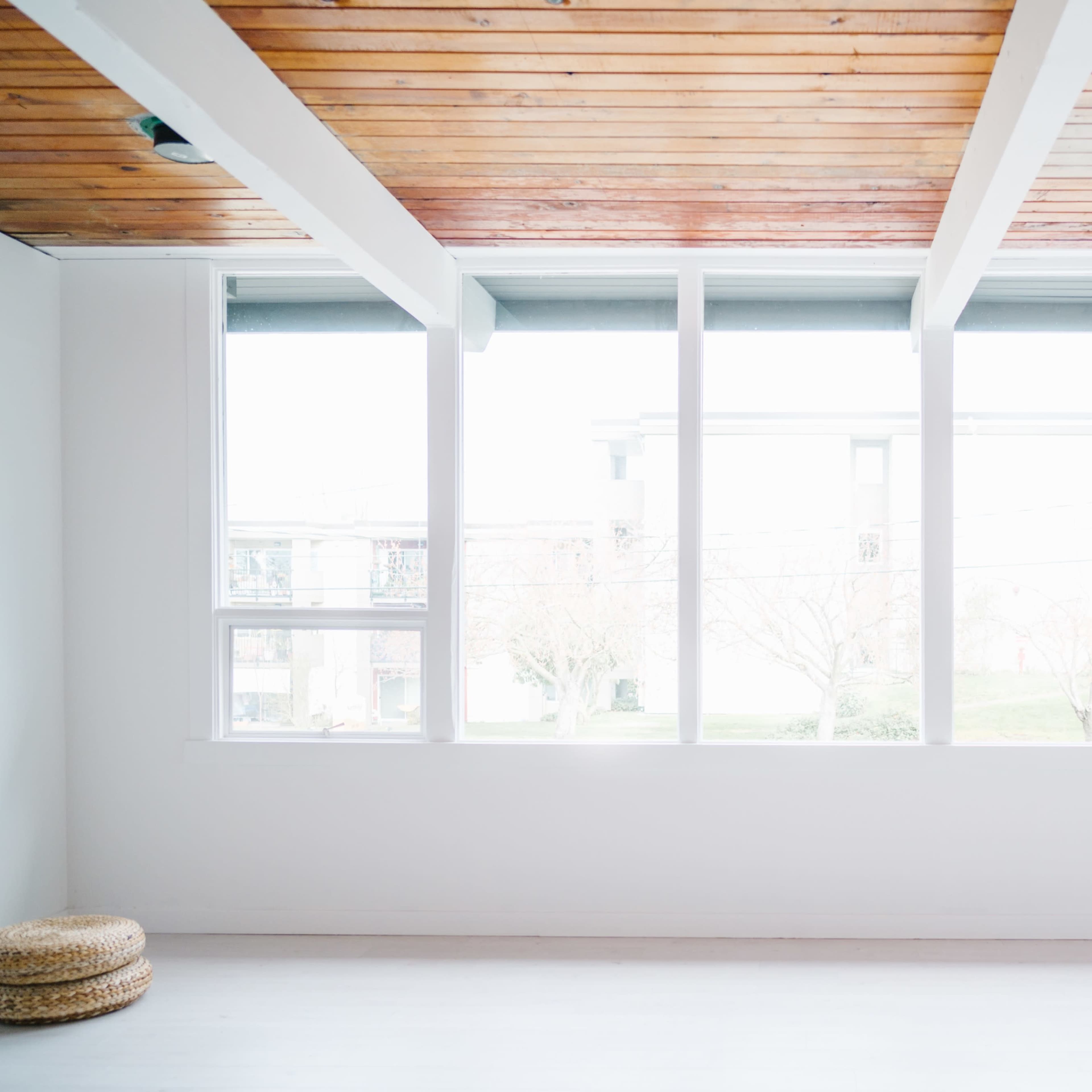 A minimalist interior room with large windows allowing natural light to fill the space, and a wooden ceiling above.