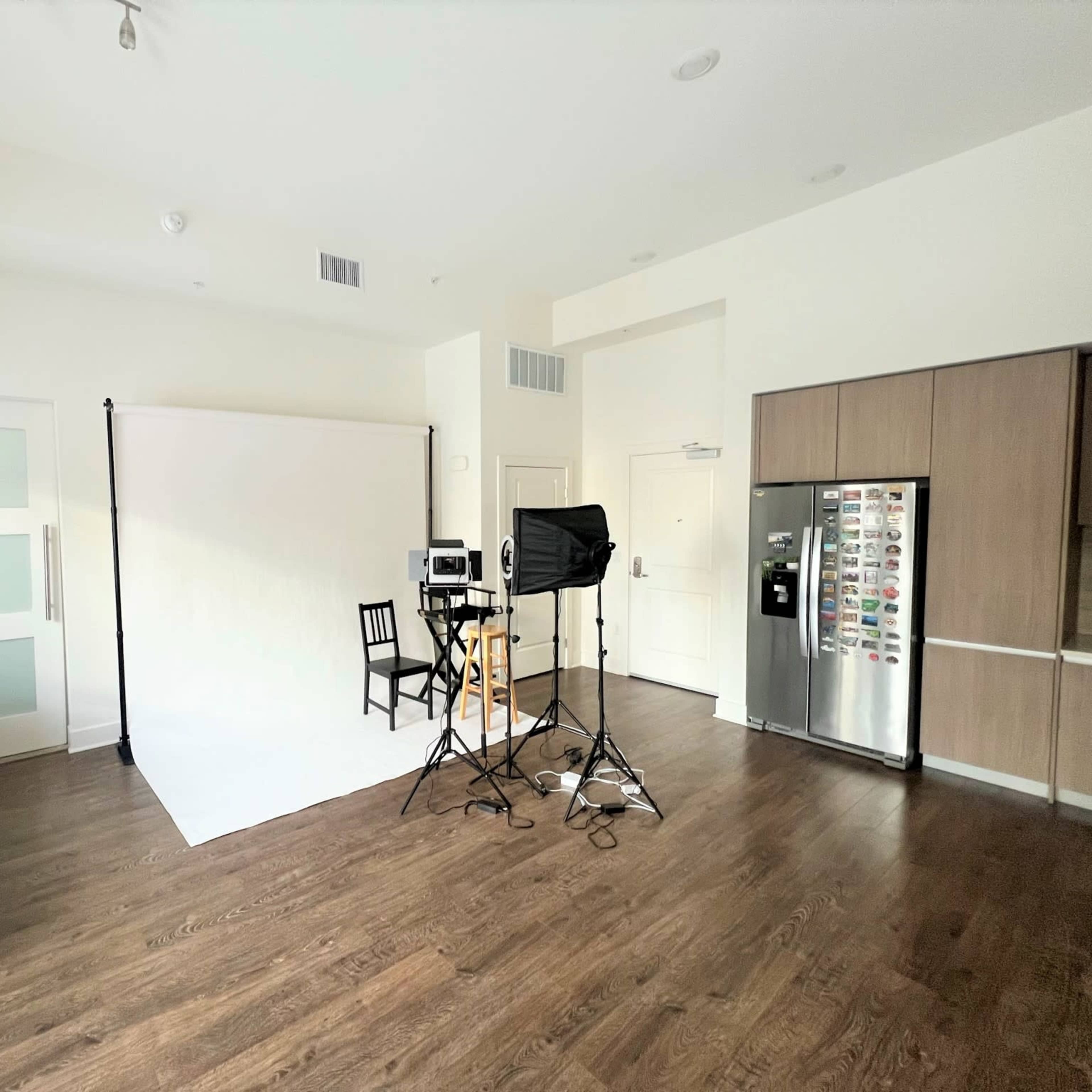 A spacious room with a white backdrop set up for photography, a chair, and lighting equipment, alongside a sofa and kitchen area with a refrigerator.