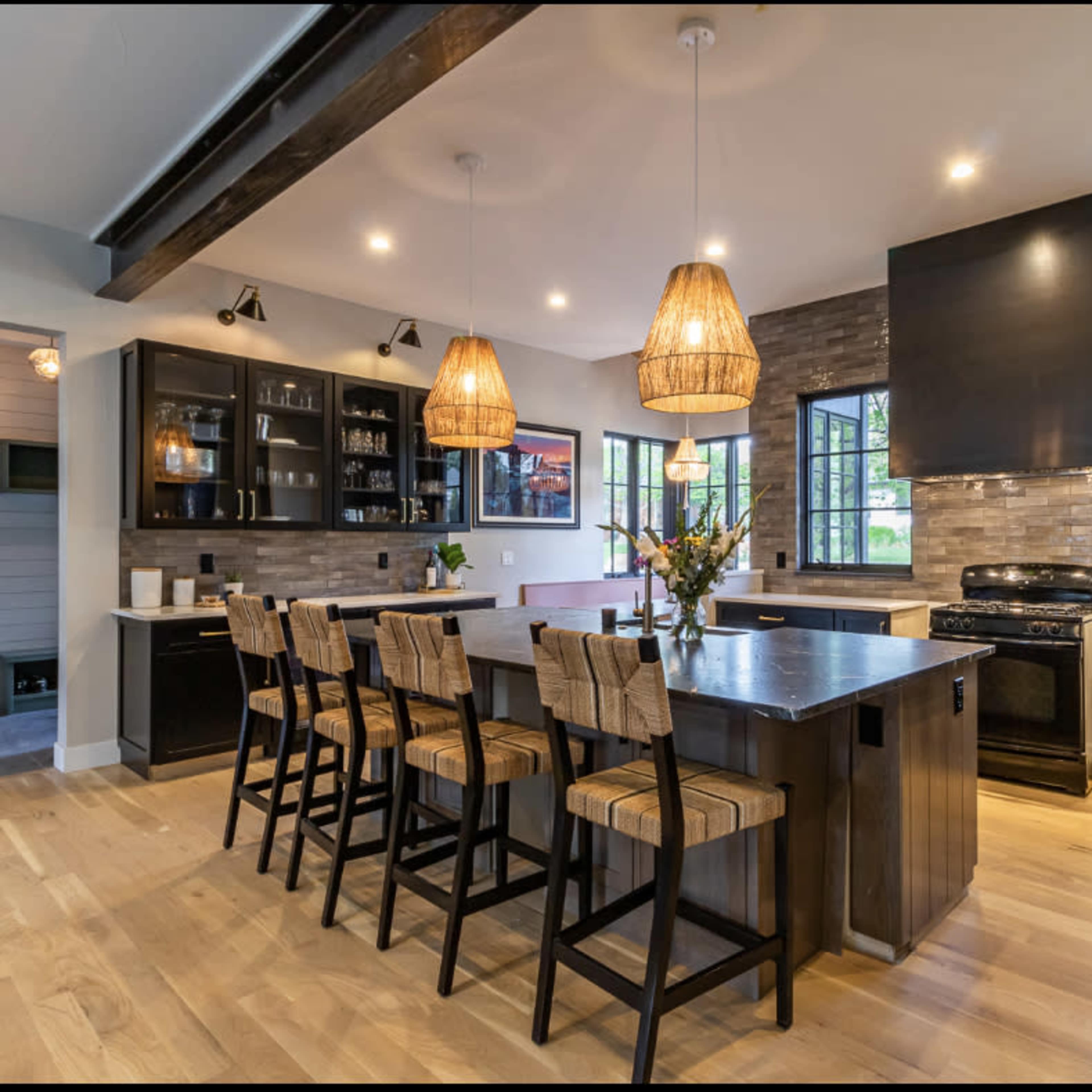 A modern kitchen features a large island with bar stools, pendant lighting, and black cabinetry, while natural light streams through multiple windows.