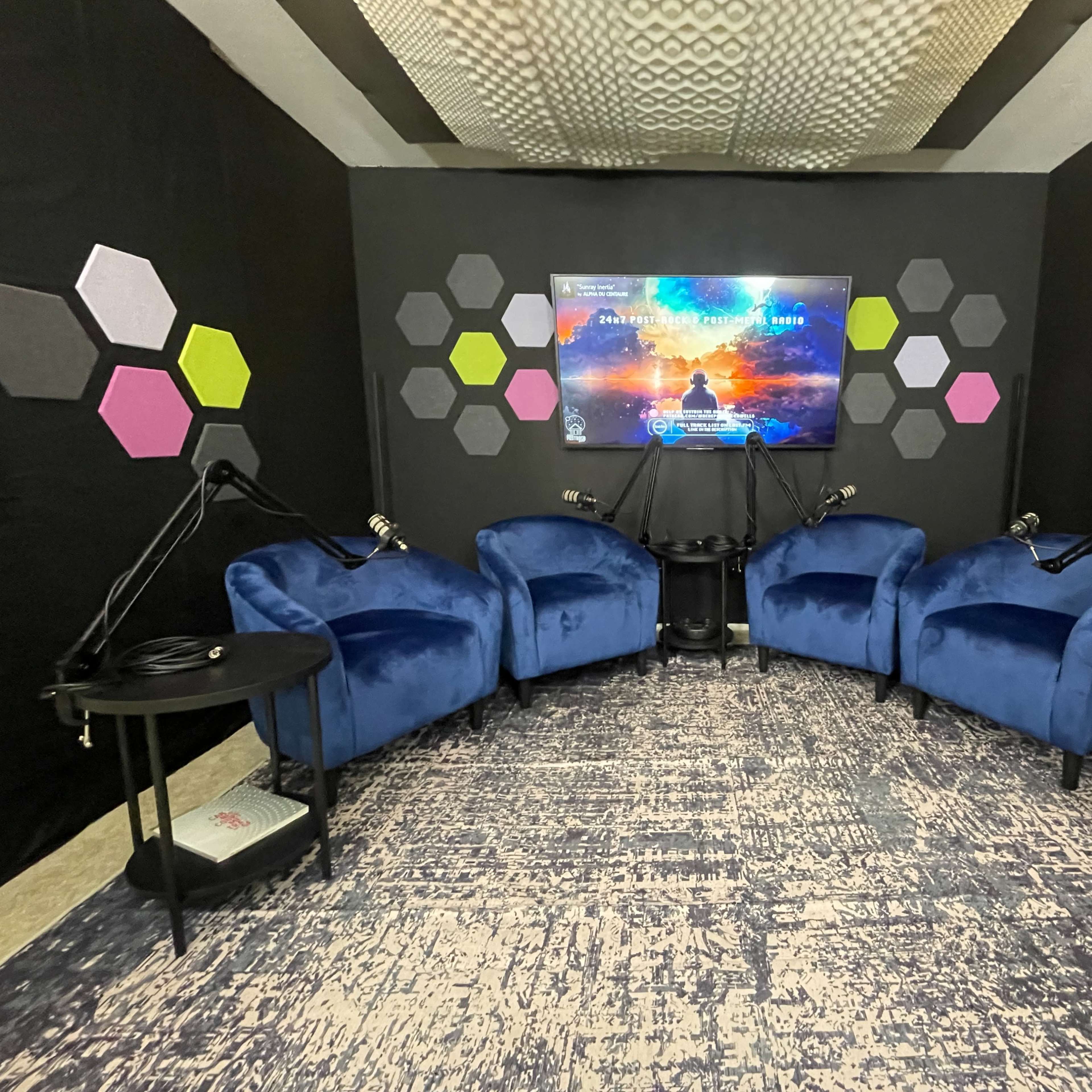 A podcast studio with four blue chairs arranged in a circular layout, facing a television screen mounted on a black wall decorated with colorful hexagonal shapes.