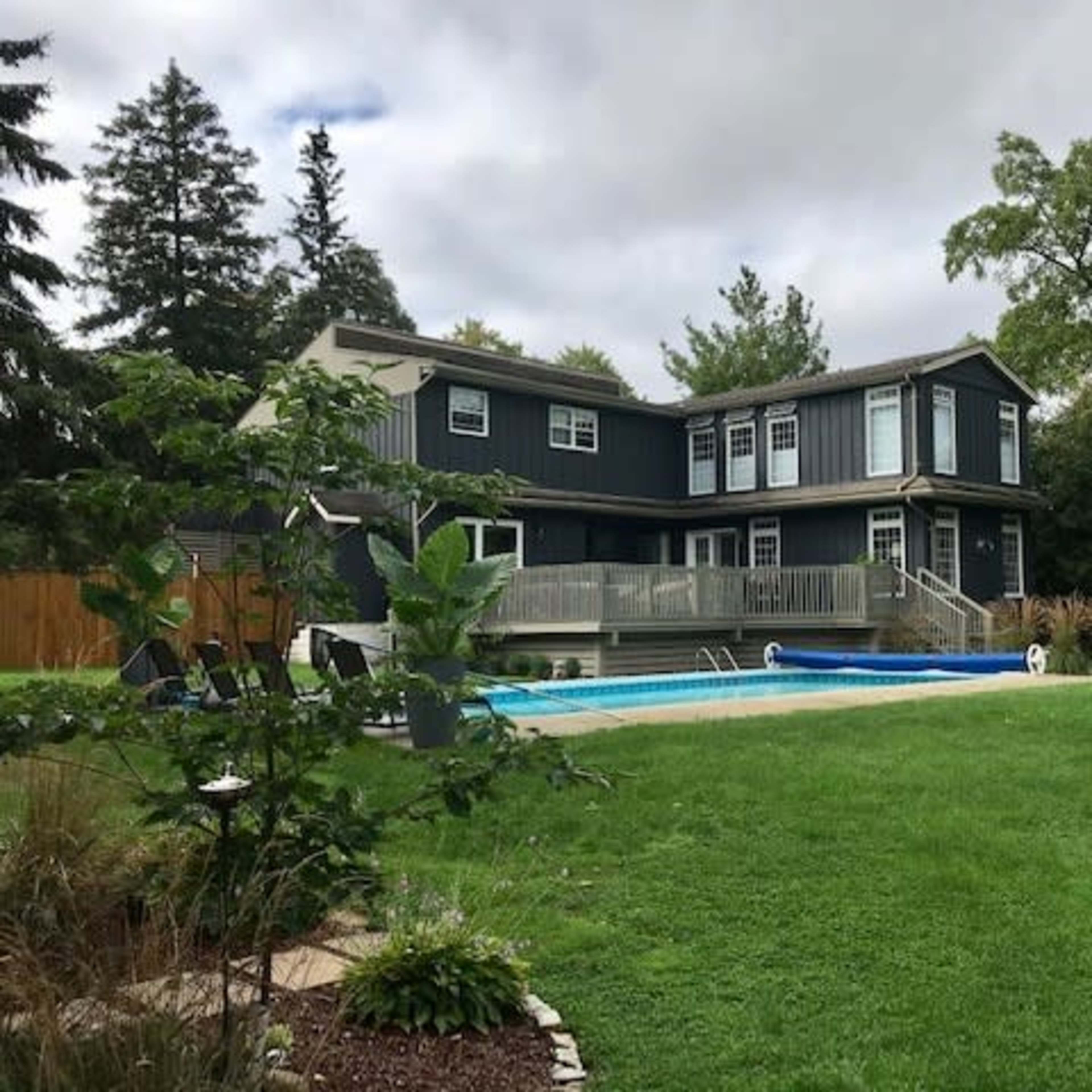 A modern two-story house with a deck overlooks a swimming pool and landscaped lawn surrounded by trees.