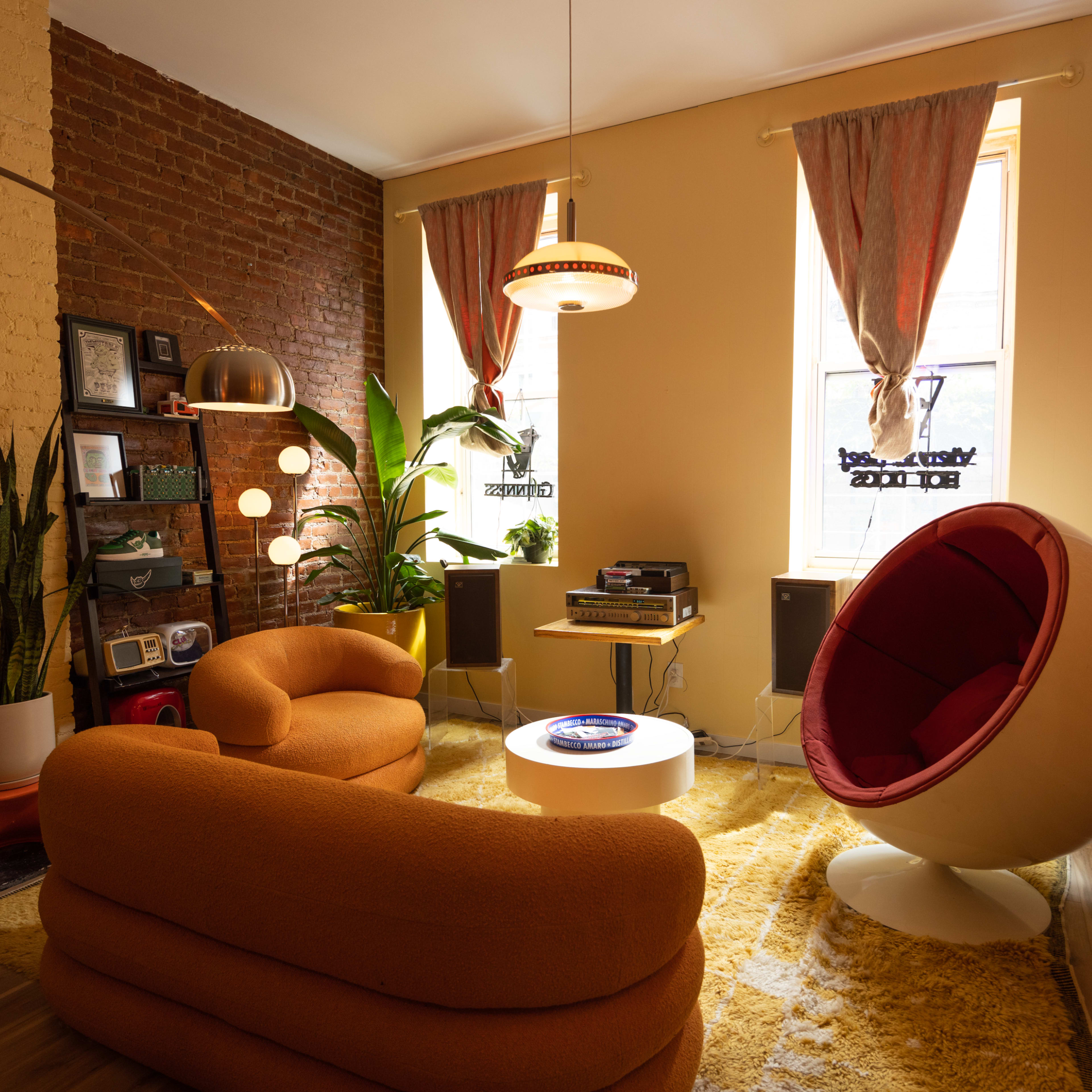 A cozy living room featuring a circular chair, two orange sofas, plants, a record player, and large windows with curtains.