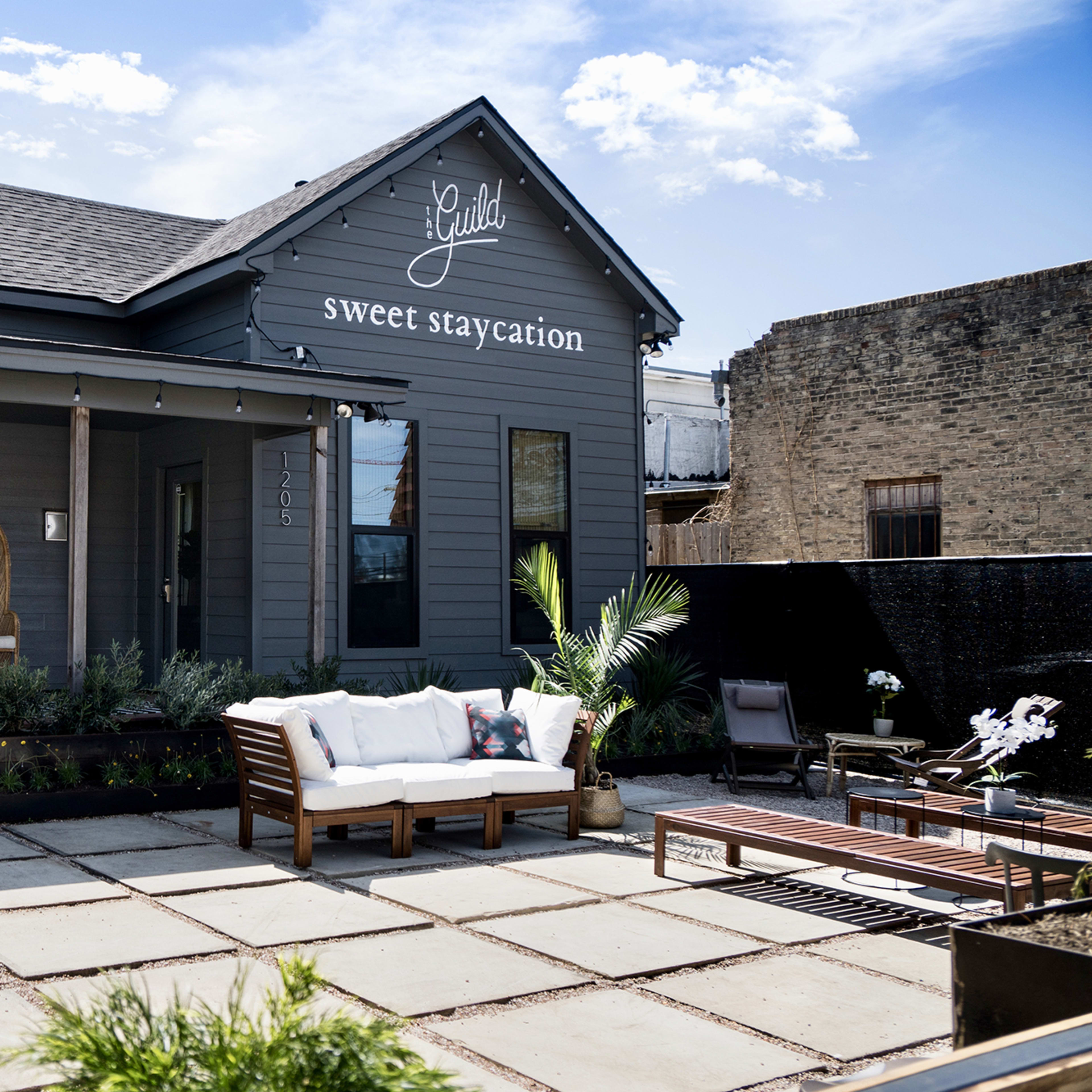 A gray house with a sign reading "sweet staycation" features a front porch with two wicker chairs and a seating area made of stone pavers.