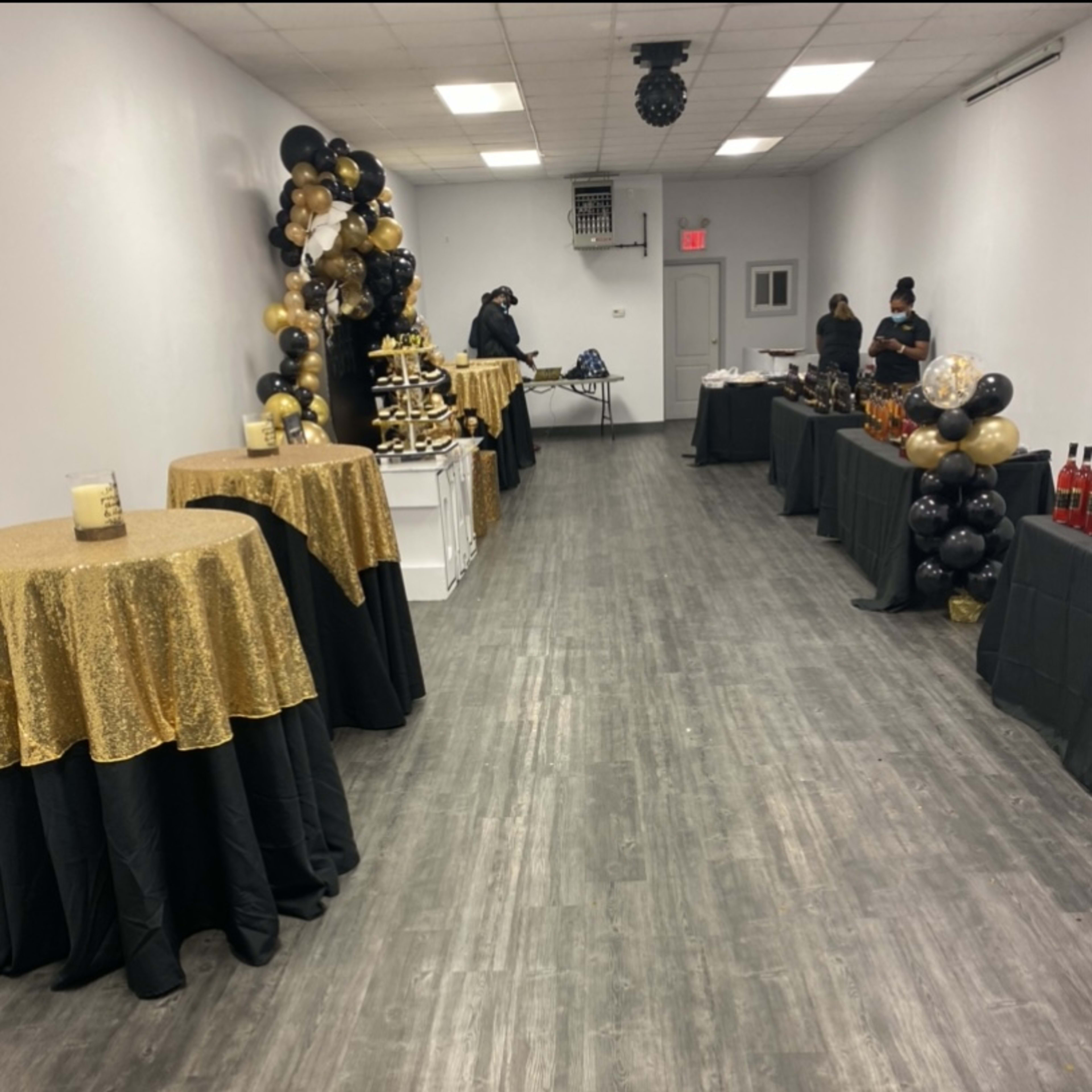 A spacious, decorated room with tables covered in gold and black cloths, set up for an event, along with a display of drinks on tables at either end.